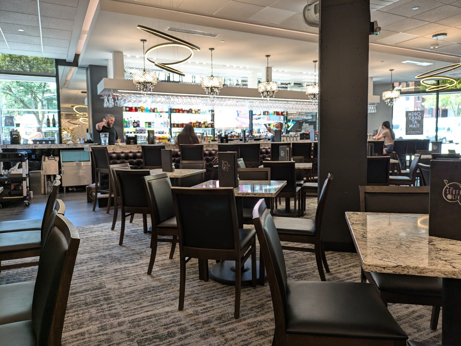 A spacious interior of The Stuffed Olive in Madison, featuring modern decor with tables and chairs, a well-stocked bar in the background, and natural light coming through large windows.
