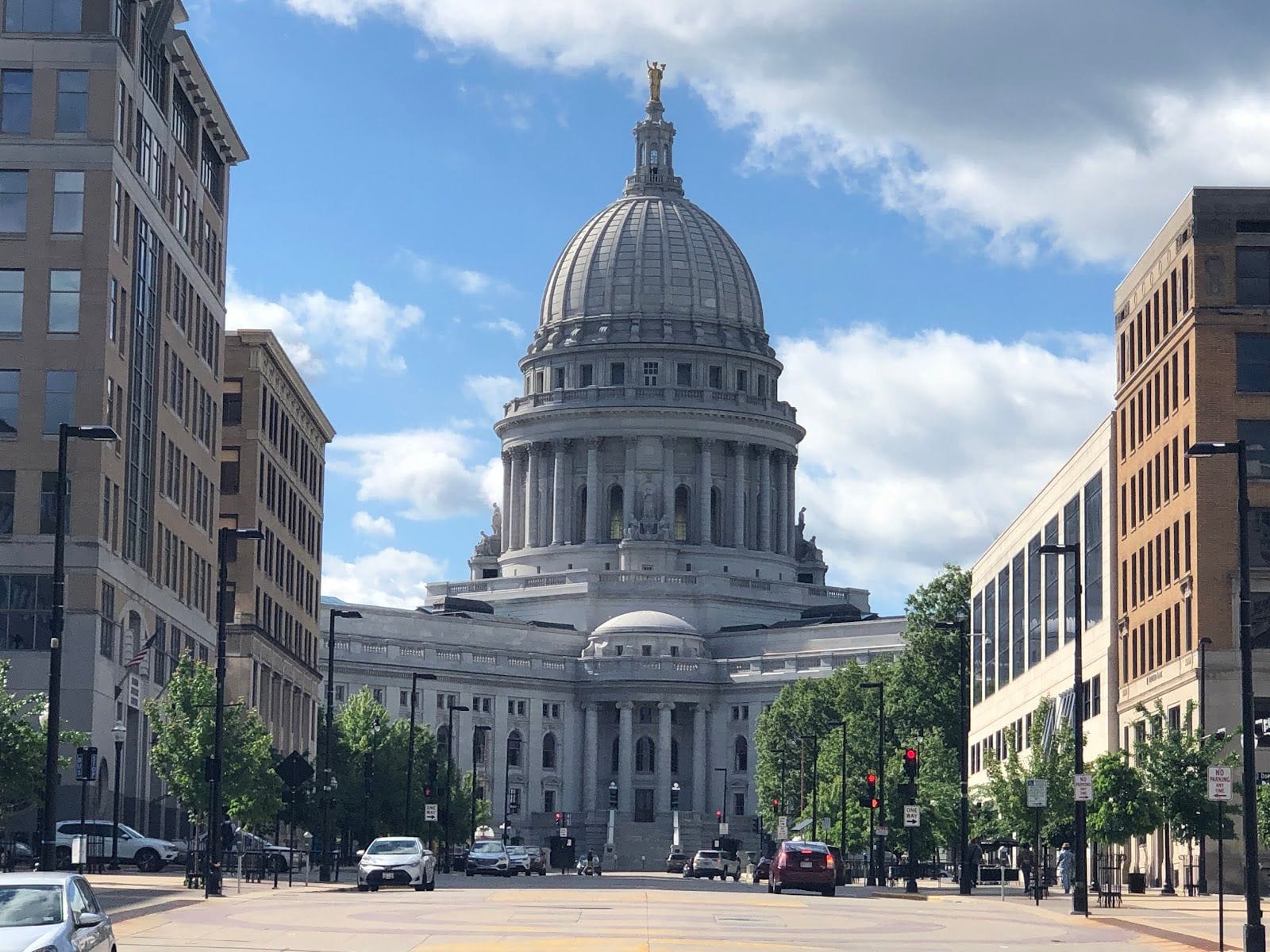 Wisconsin State Capitol stands tall under a blue sky, framed by city buildings and greenery.