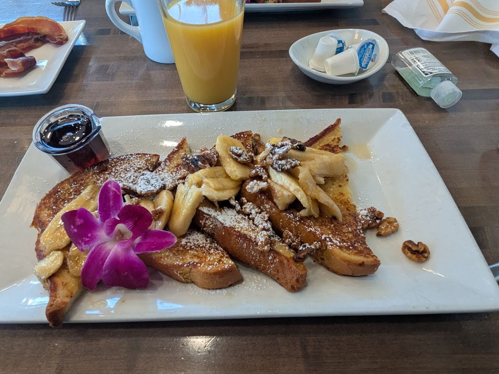 French toast topped with bananas, walnuts, powdered sugar, a purple flower, and syrup on a white plate.