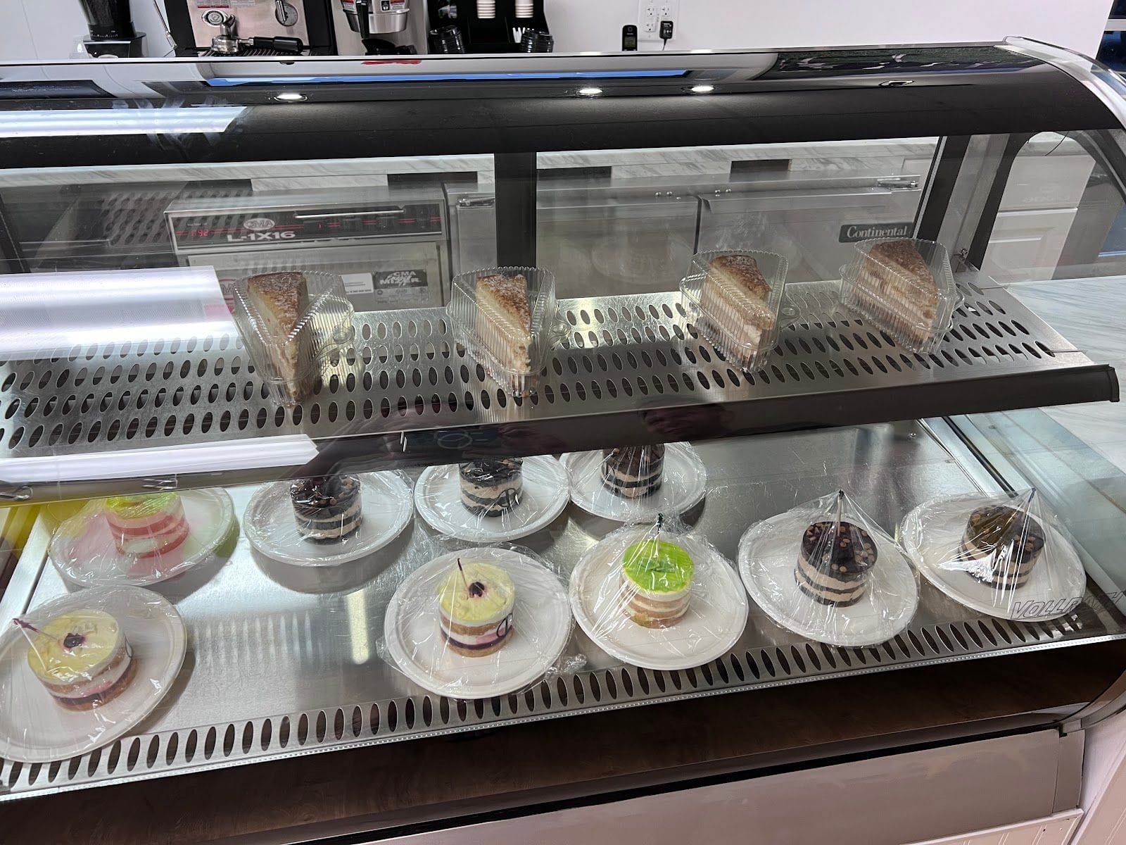 Pastry display case with assorted cakes and desserts, some wrapped in plastic, in a bright shop setting.