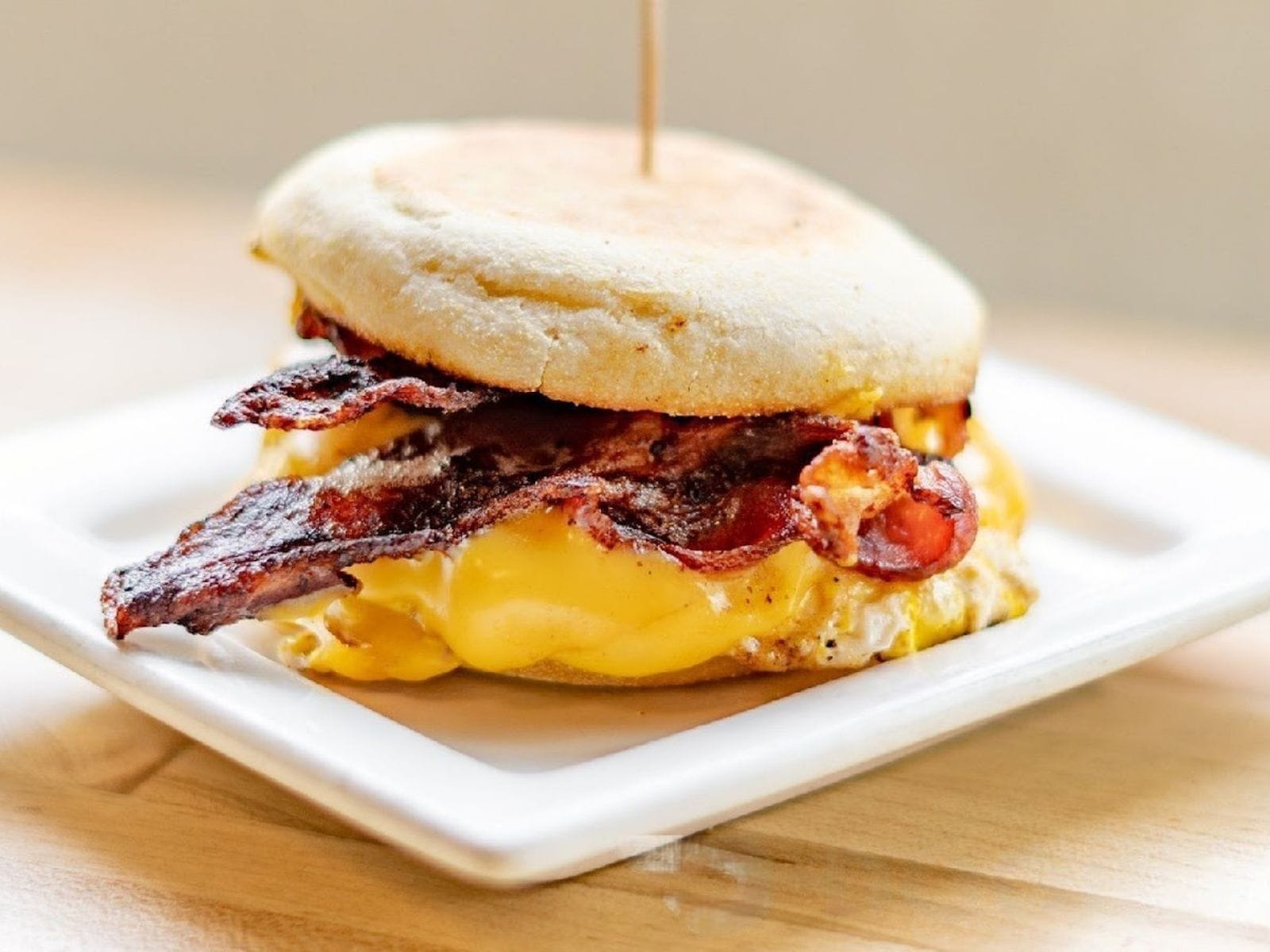Bacon, egg, and melted cheese breakfast sandwich on an English muffin served on a square white plate.