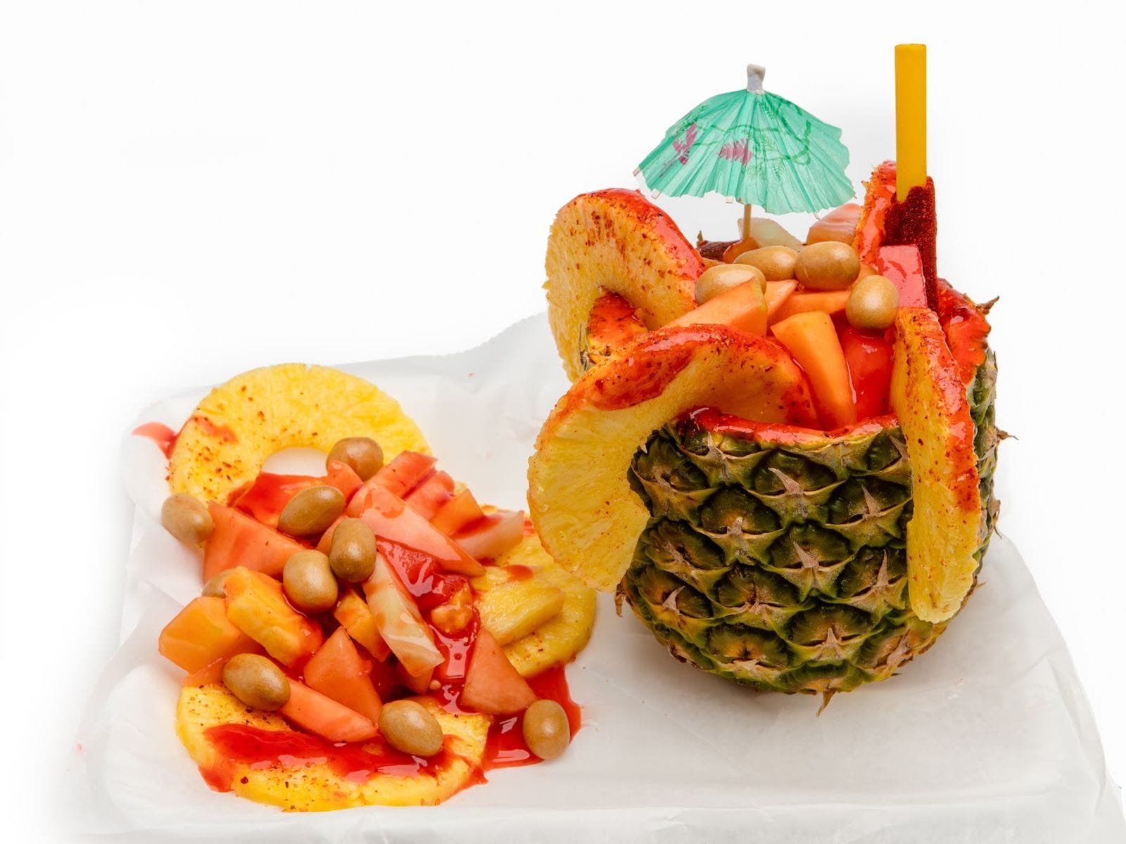 Pineapple bowl filled with fruit, chamoy sauce, candy, and a paper umbrella, with extra fruit on the side.
