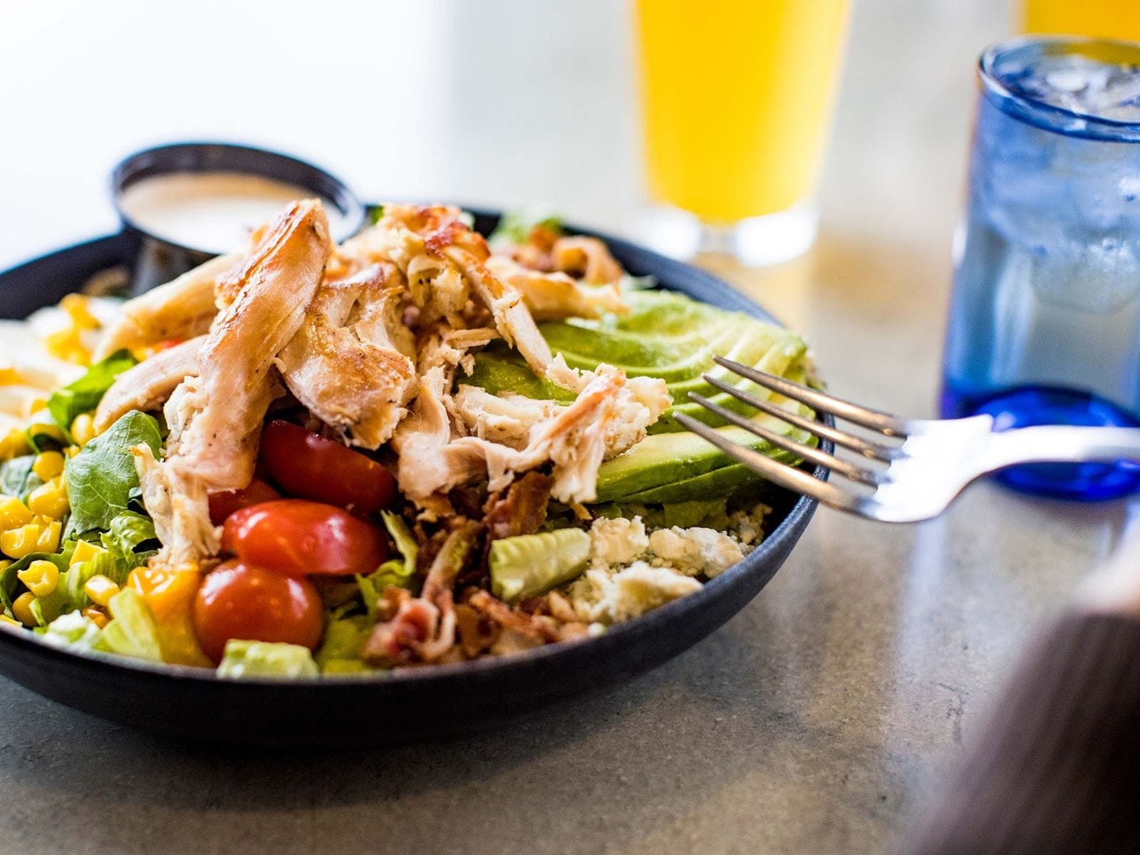 Fresh salad with grilled chicken, avocado, cherry tomatoes, corn, and dressing at a restaurant table.