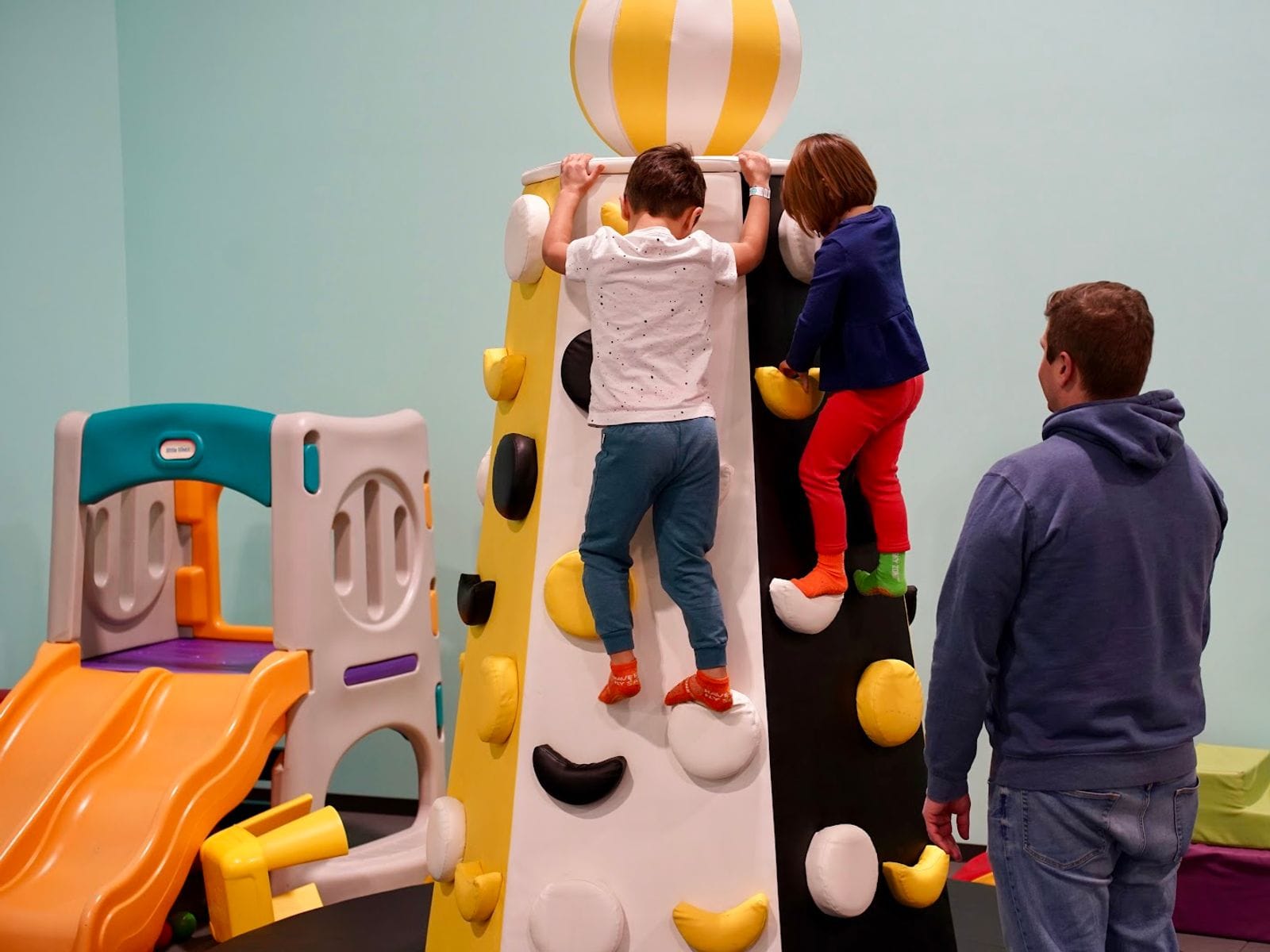 Two kids climb a colorful indoor play structure while an adult supervises at Wisco Kidz Place LLC.