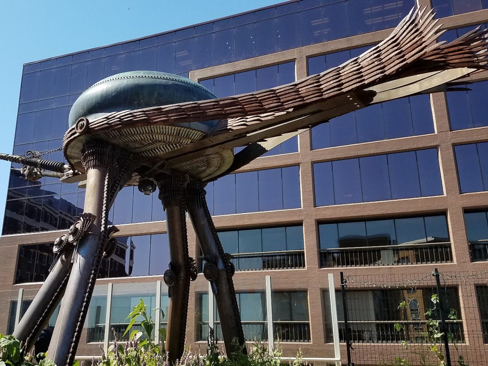 Large metal insect sculpture with long legs and wings outside Madison Children’s Museum, modern building behind.