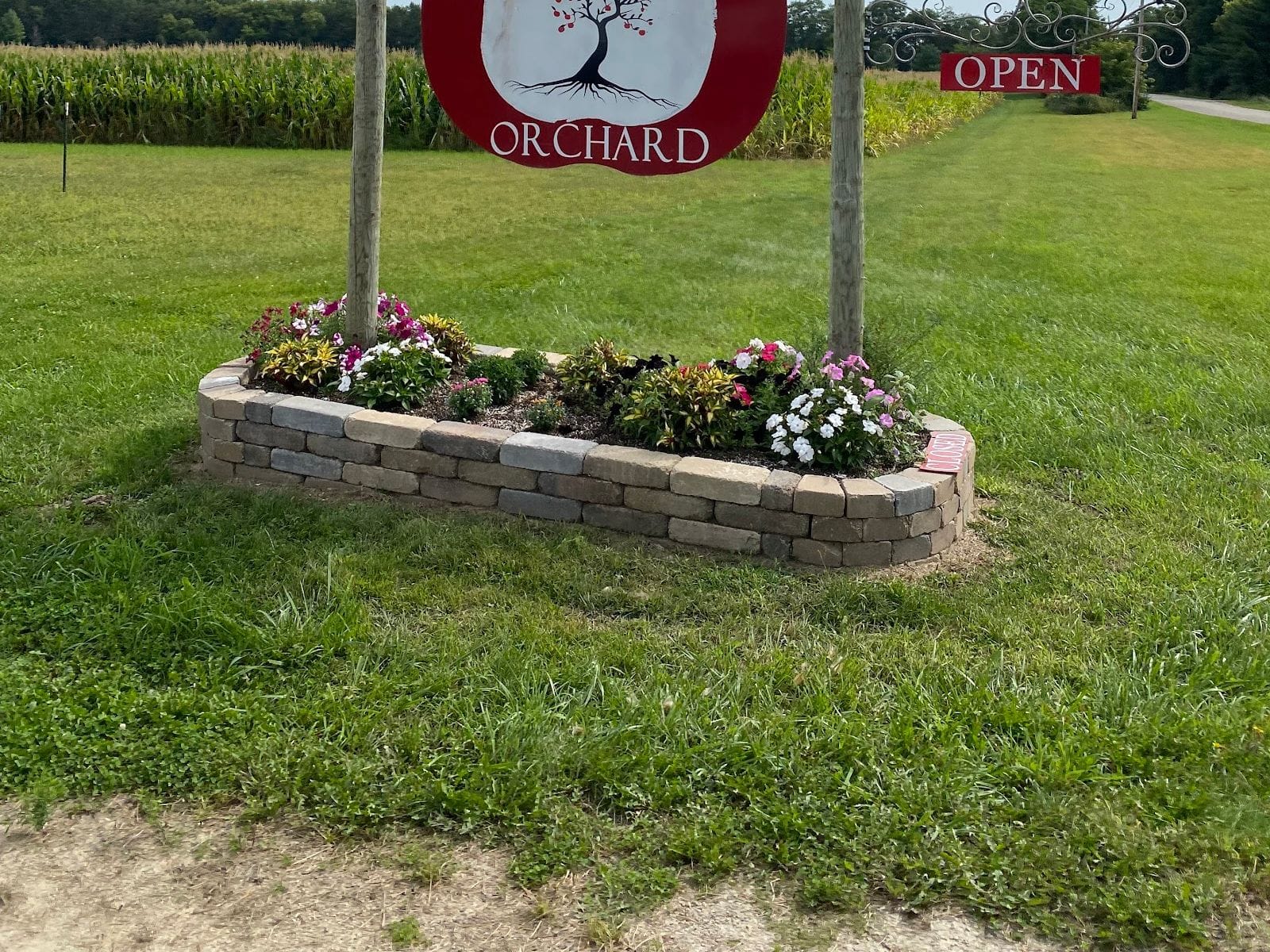 Wooden Alpine Ridge Orchard sign with flowers in a stone planter, surrounded by green grass and a cornfield.