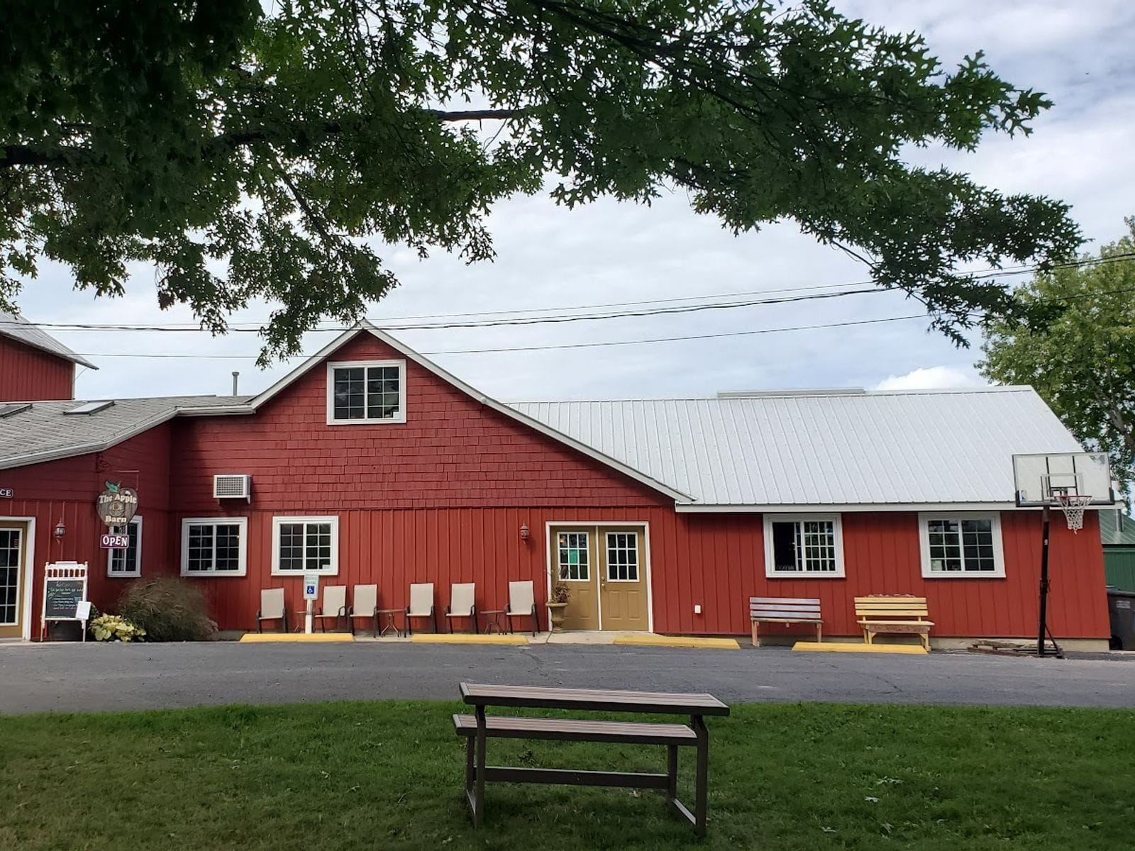 Red barn building with white trim, benches, and picnic table under leafy trees at Apple Barn Orchard & Winery.