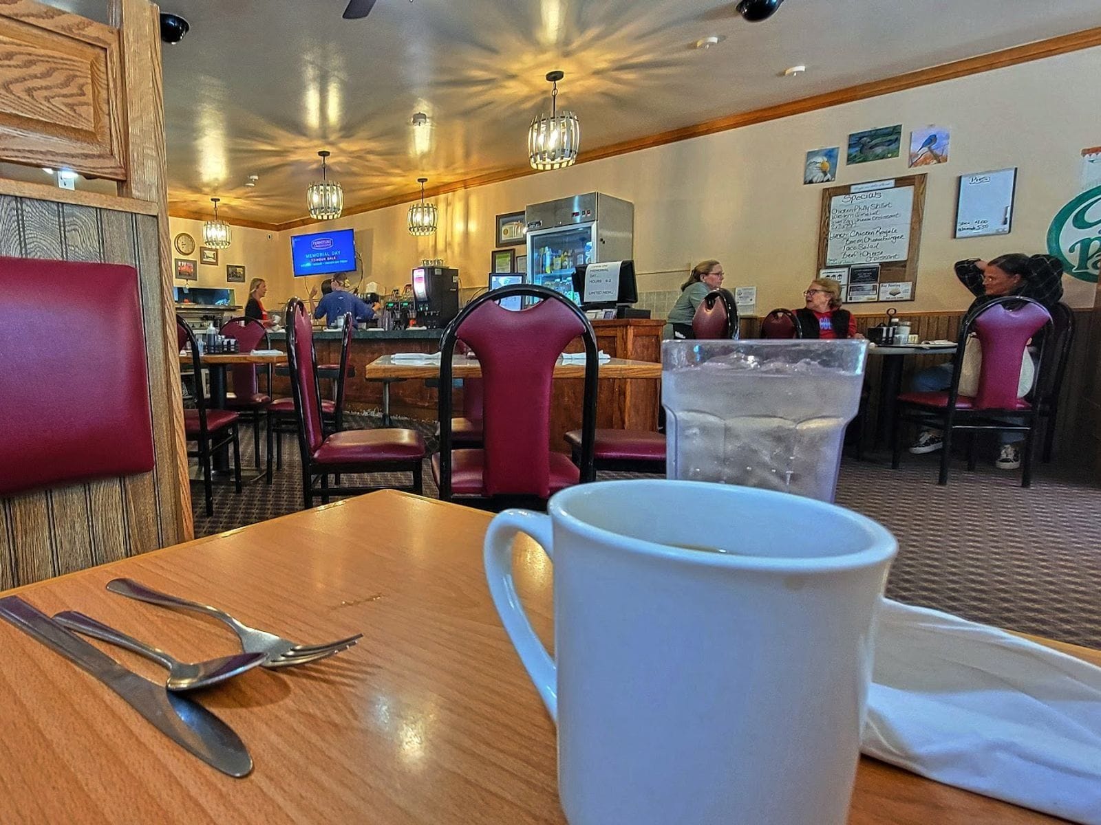 Coffee mug and water glass on a table in a cozy diner with red chairs and people dining in the background.
