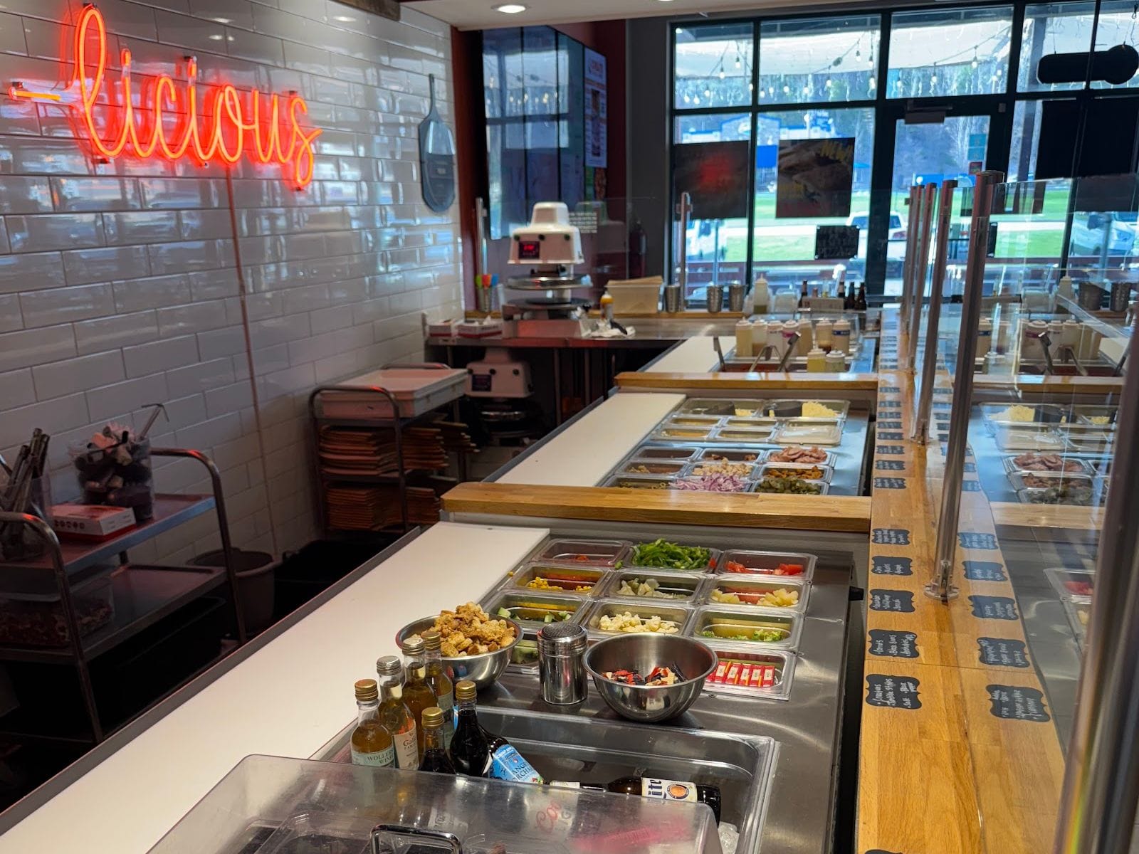 Pizza toppings bar with fresh ingredients at Dells Pizza Lab, neon sign and bright windows in background.