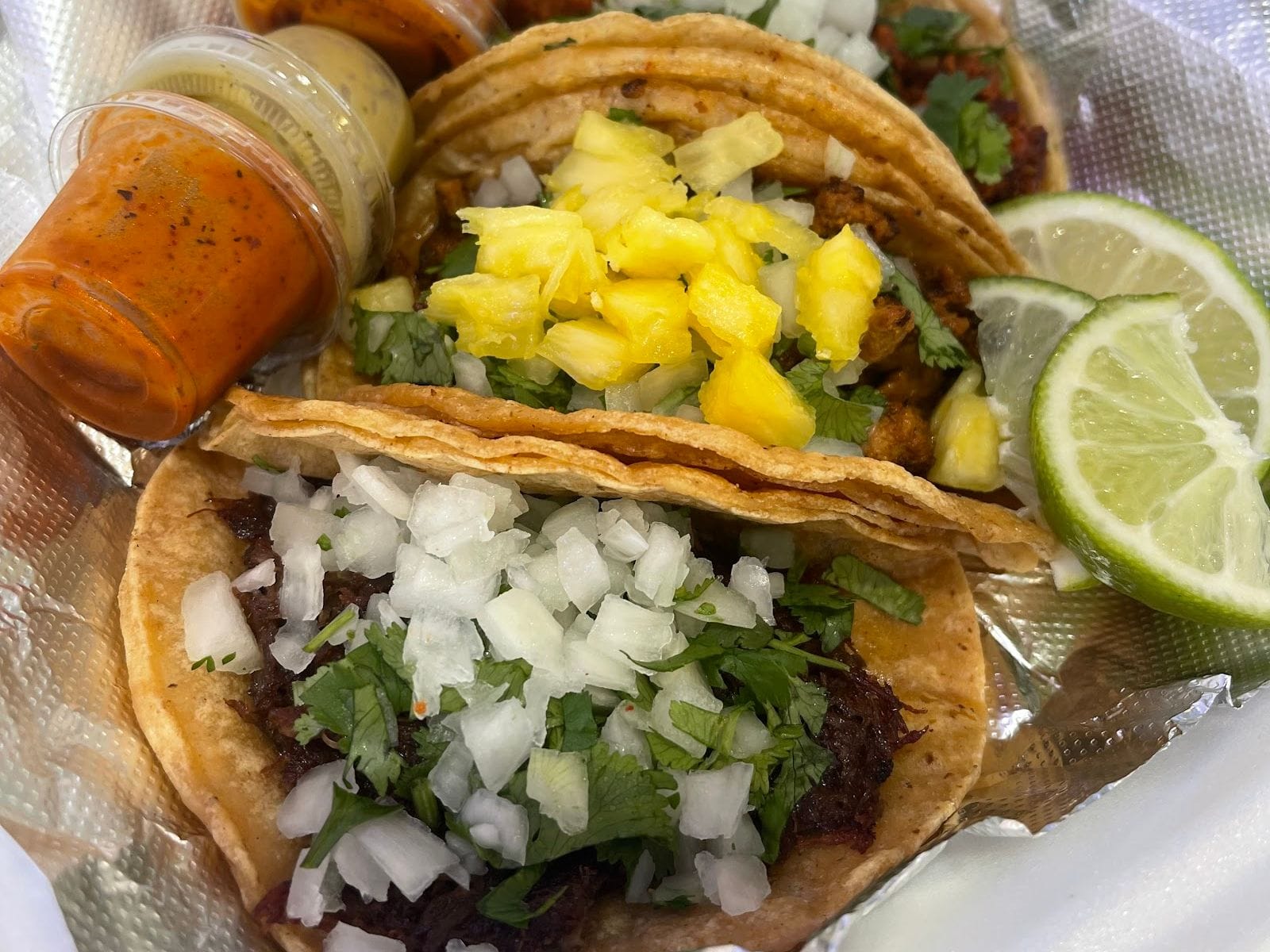 Two tacos topped with onions, cilantro, pineapple, lime wedges, and sauces in a takeout container.