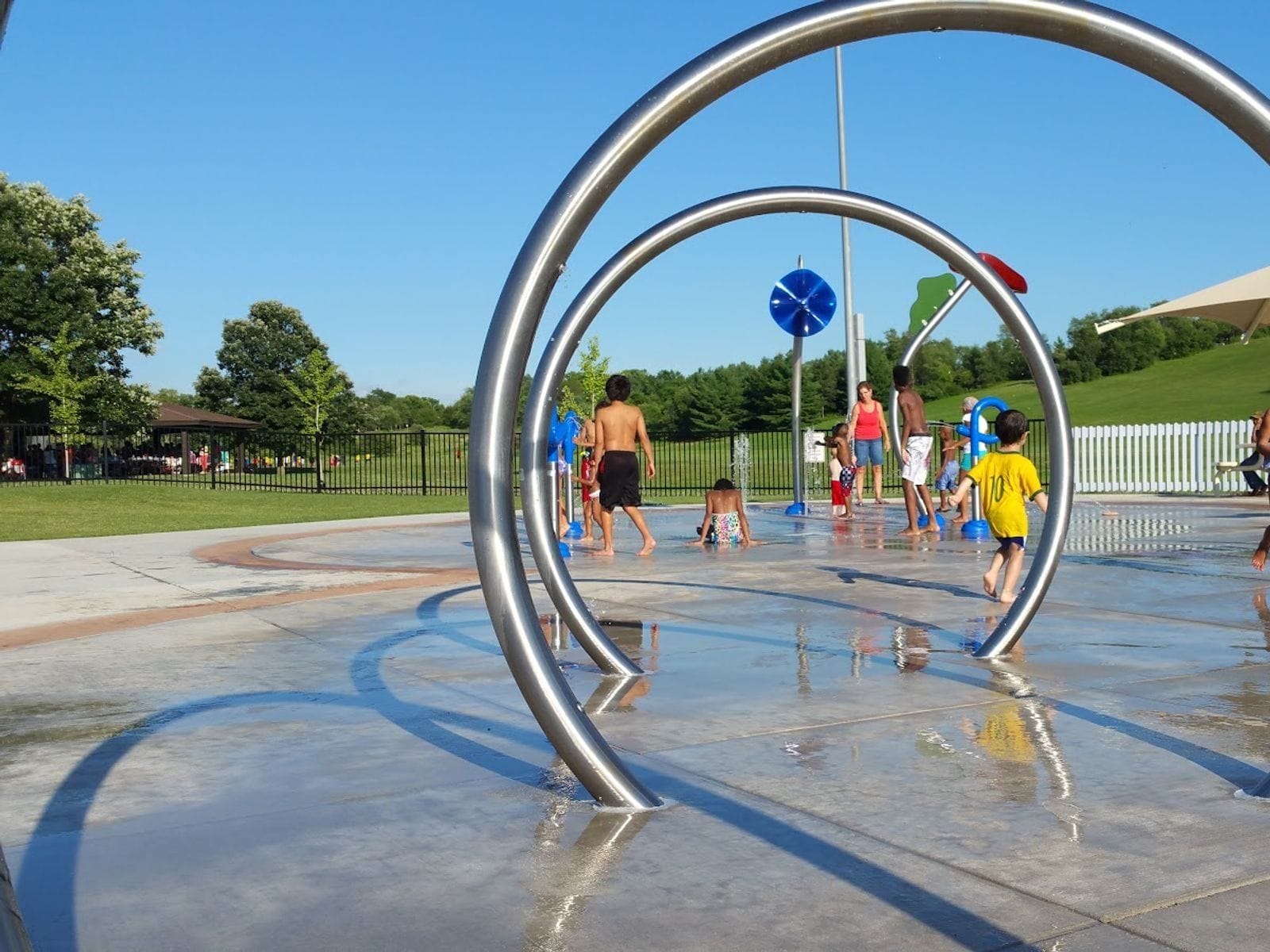 Children play in a splash pad with metal arches and water jets at Elver Park on a sunny day.