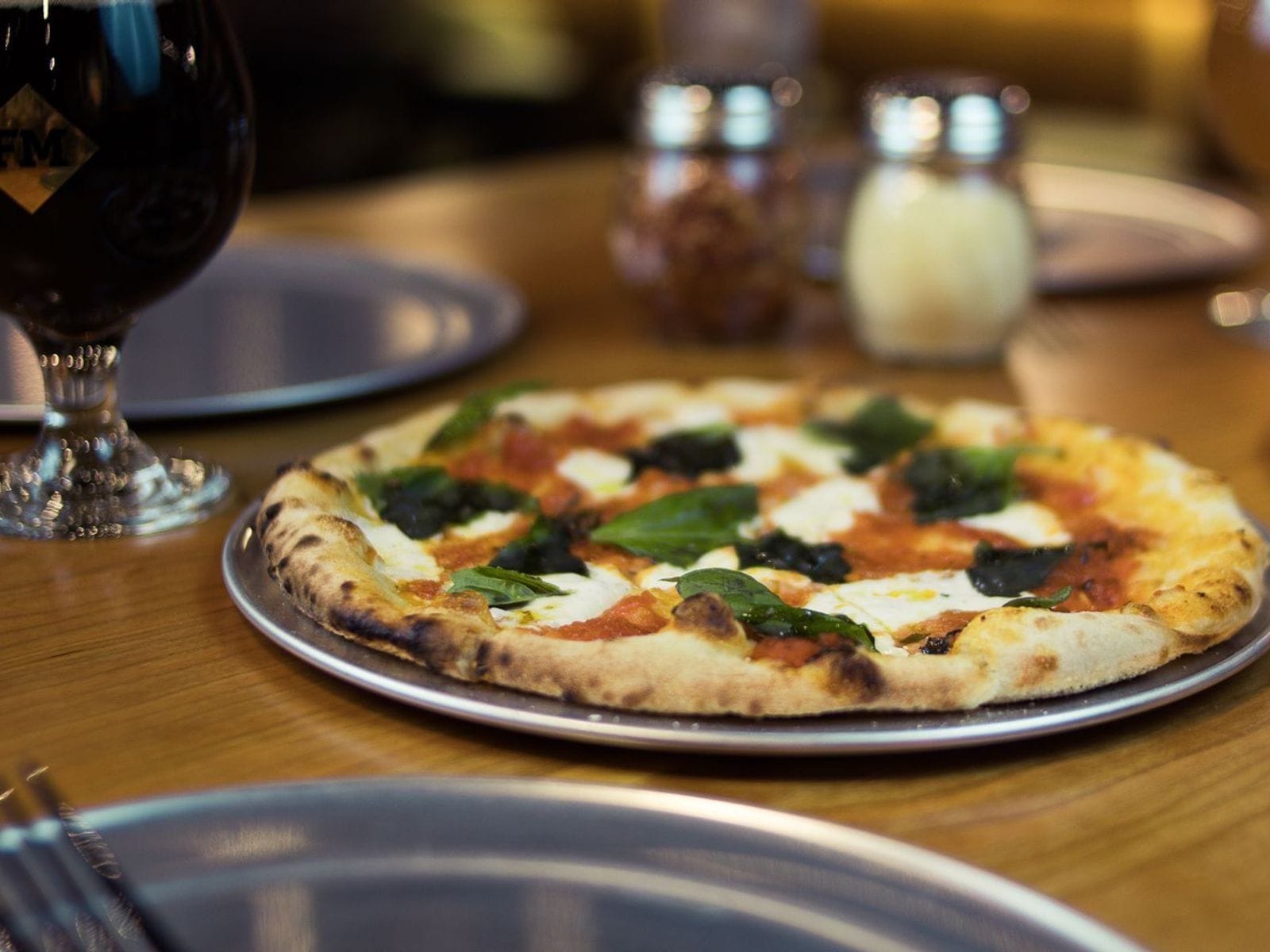 Fresh margherita pizza with basil on a metal tray beside a glass of dark beer on a wooden table.