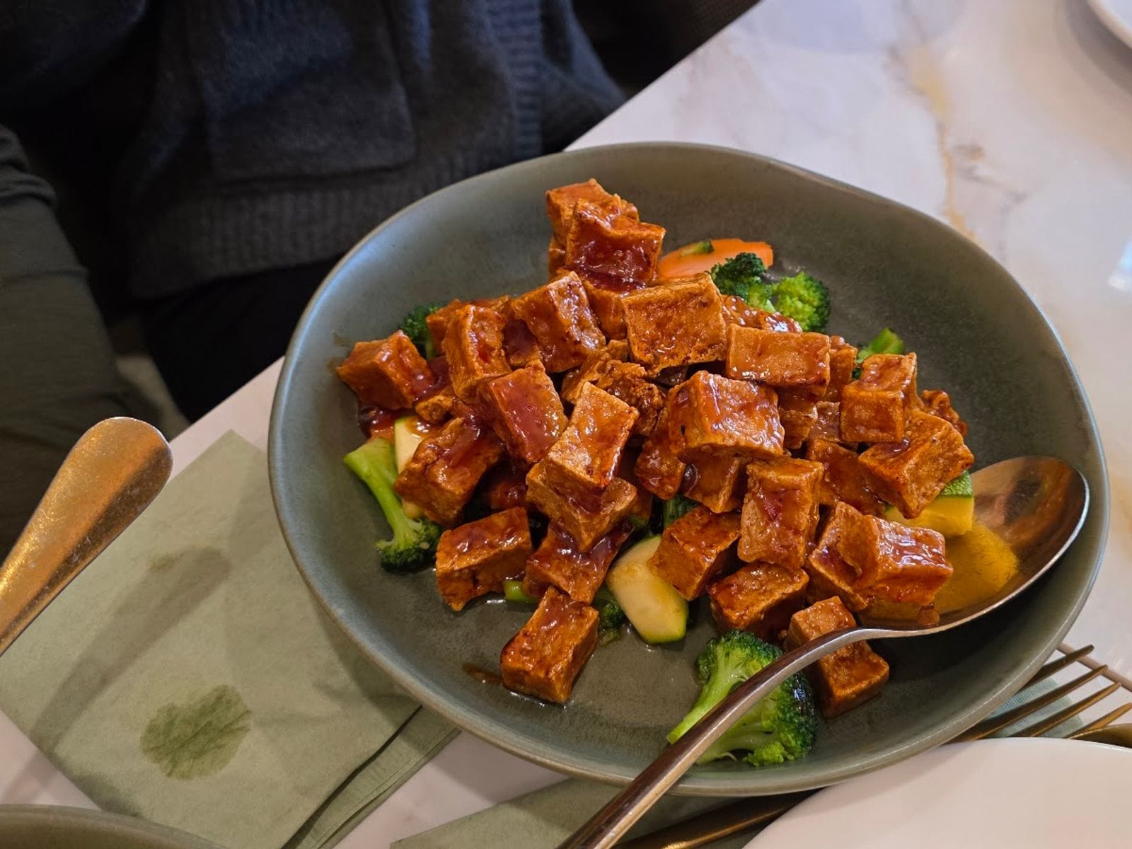 Glazed tofu cubes served over steamed broccoli and vegetables in a dark bowl at a restaurant table.