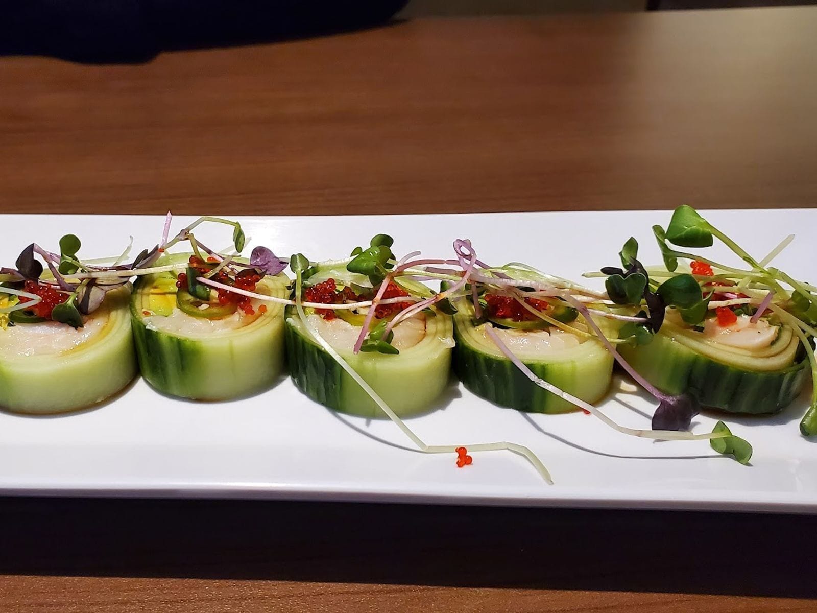 Six cucumber-wrapped sushi rolls garnished with microgreens and fish roe on a white rectangular plate.