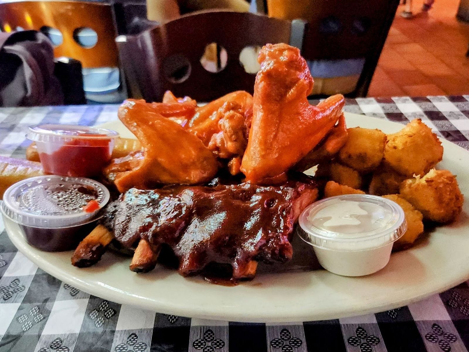 Plate of saucy ribs, buffalo wings, fried cheese curds, and dipping sauces on a checkered tablecloth.