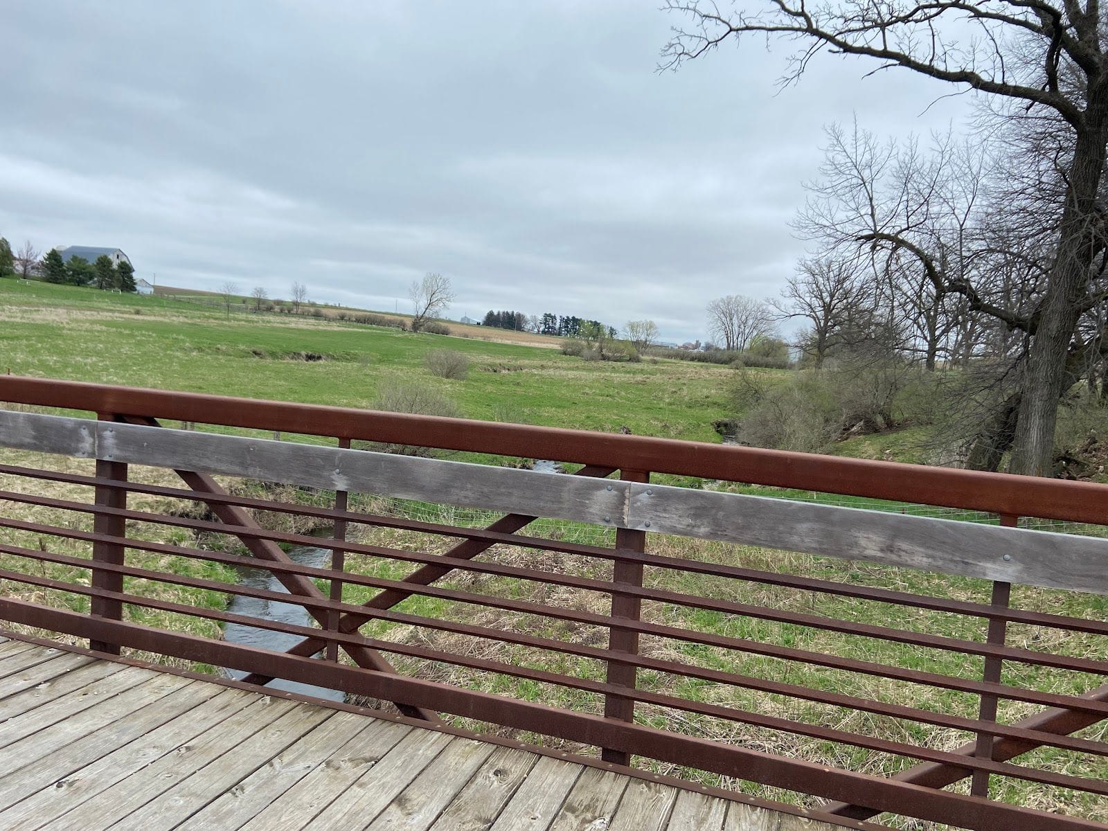 Wooden bridge over a small creek with open grassy fields and leafless trees under a cloudy sky.
