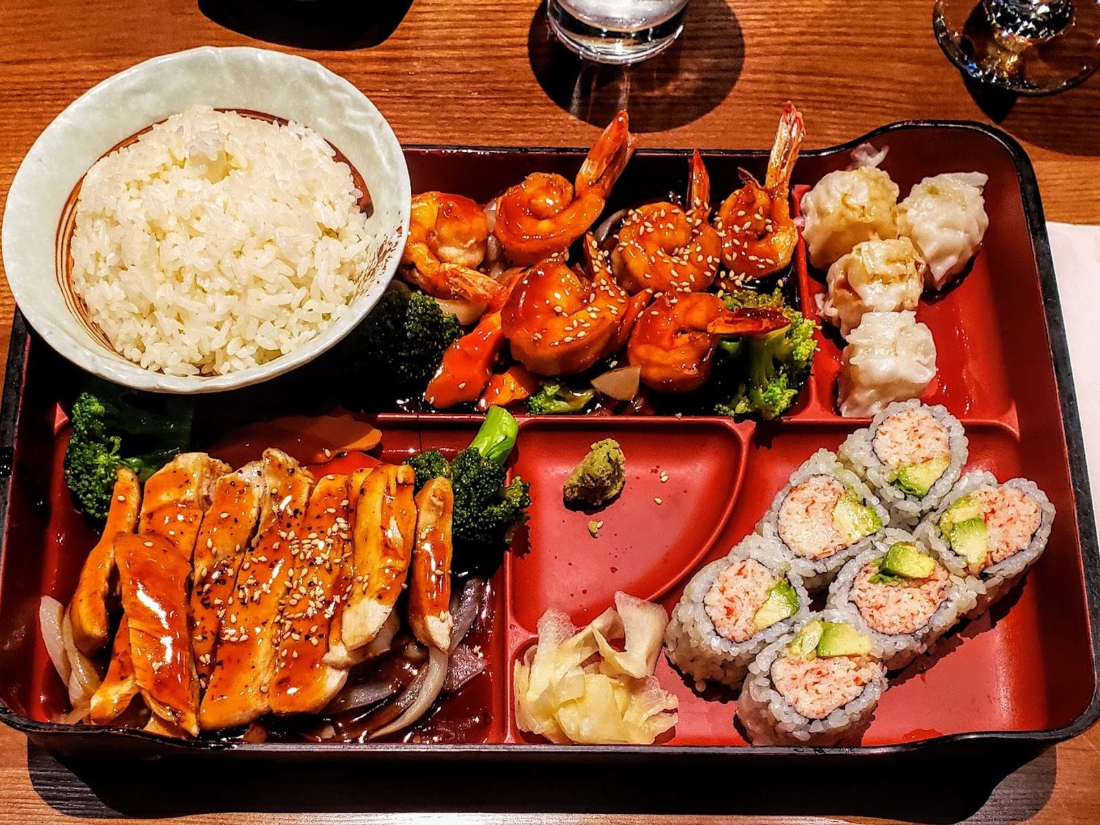 Colorful bento box with sushi rolls, shrimp, chicken teriyaki, shumai, broccoli, and white rice.