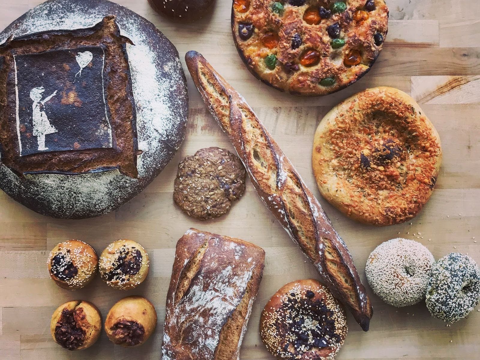 Assorted artisan breads and rolls on a wooden table, including a loaf with a stencil art crust.