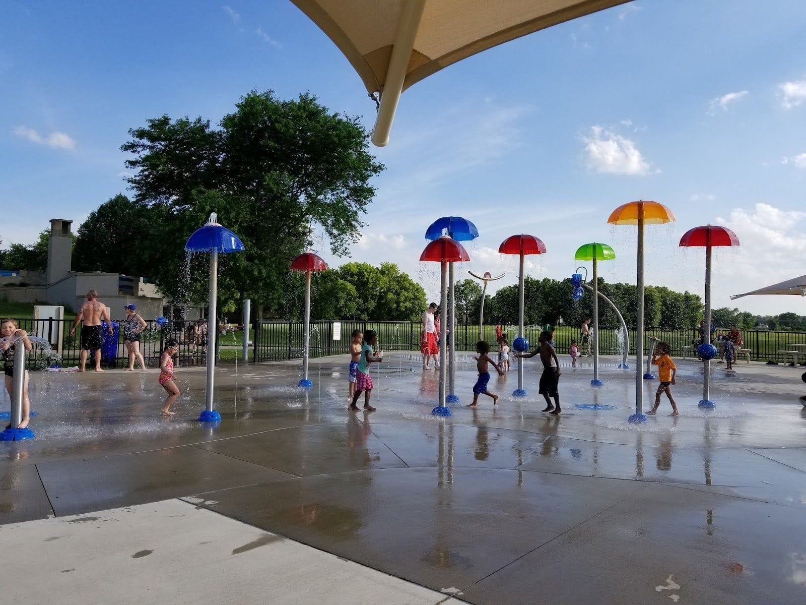 Children playing under colorful water umbrellas at a splash pad on a sunny day.