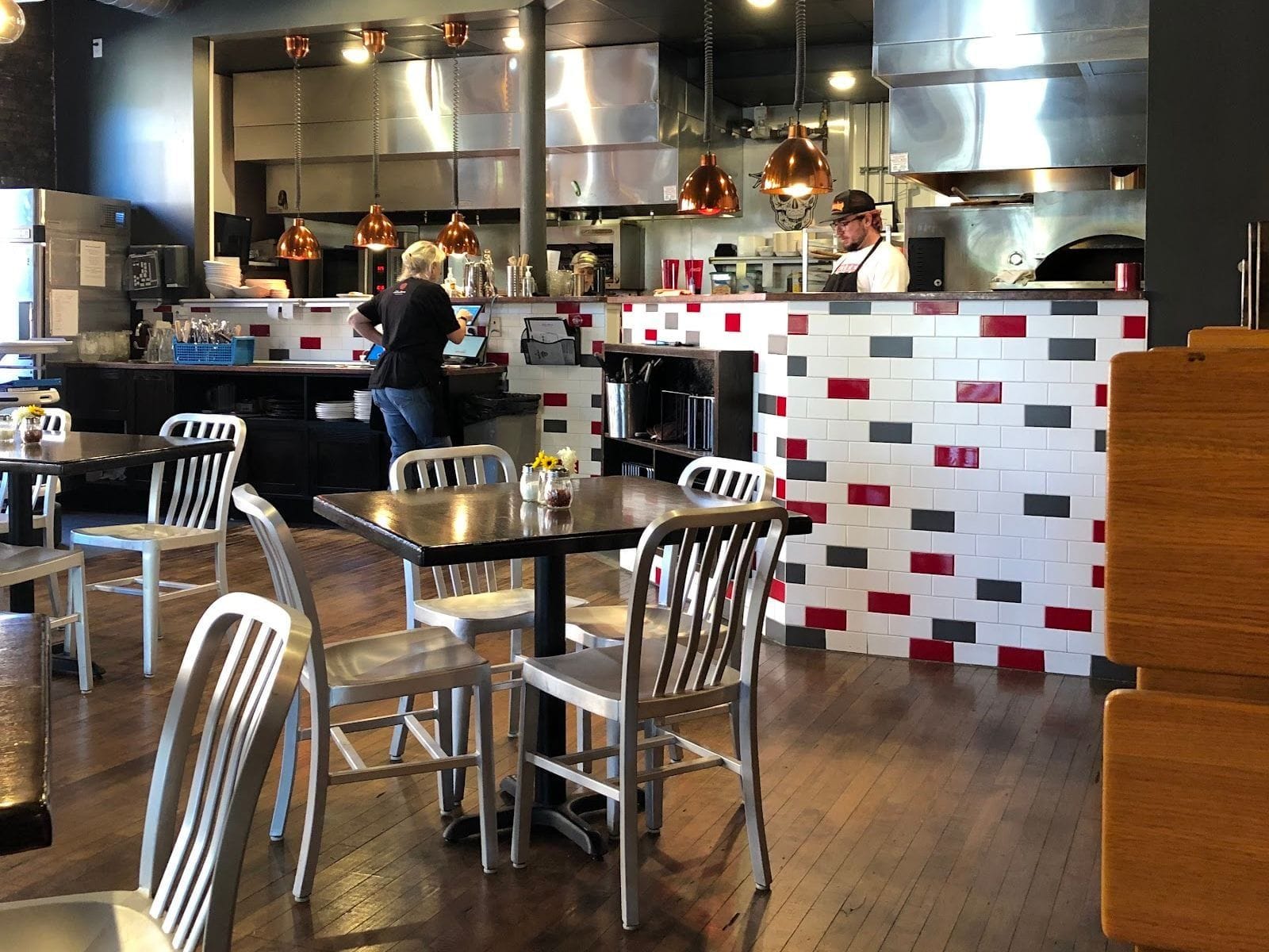 Modern pizza restaurant interior with metal chairs, wood tables, and a tiled counter in red, gray, and white.