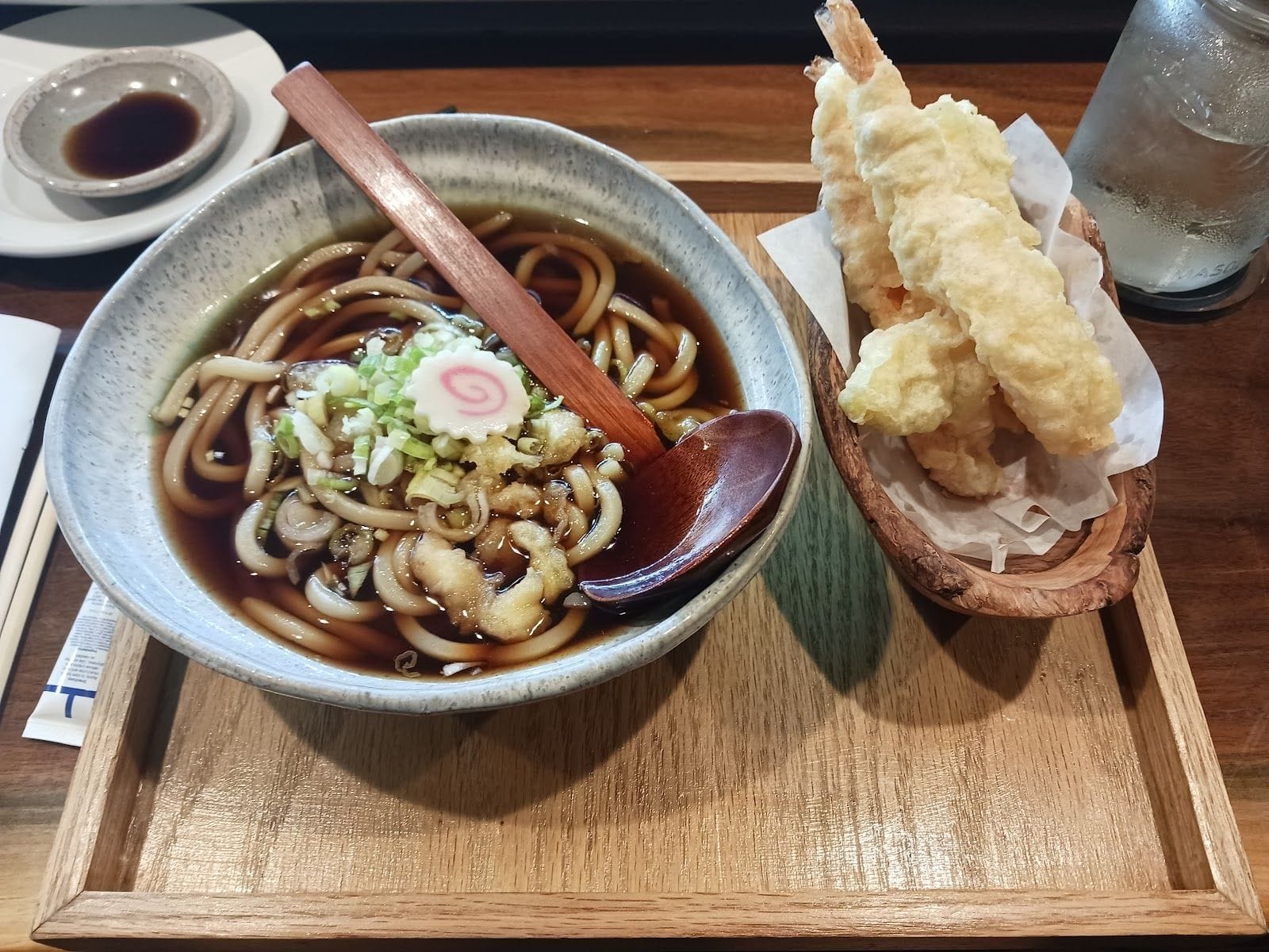 Bowl of udon noodle soup with tempura shrimp on a wooden tray at a Japanese restaurant.