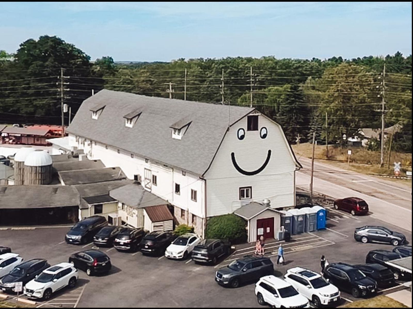Large white barn with a giant smiley face, surrounded by parked cars and trees in the background.