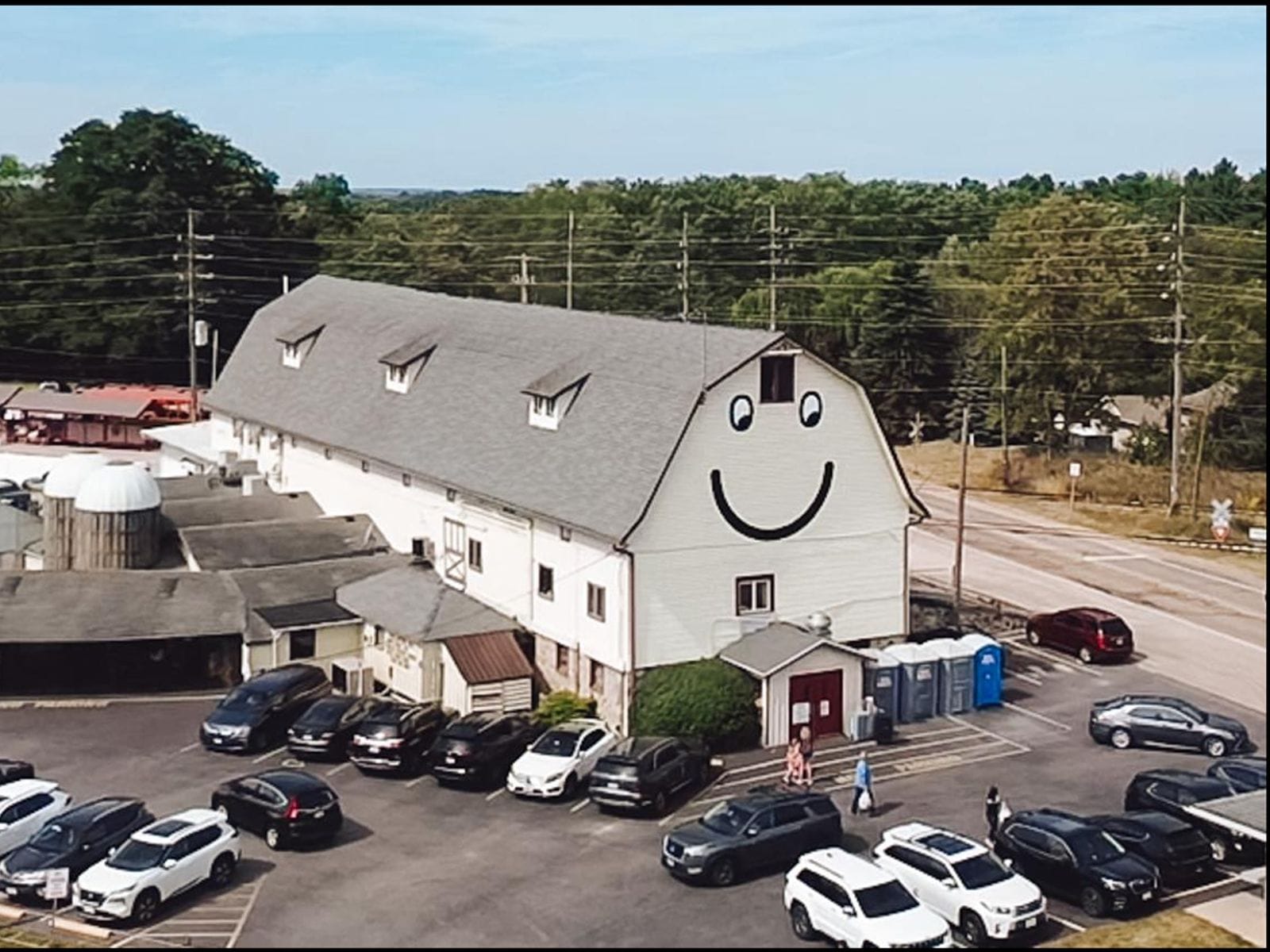 Large barn with a giant smiley face on its front, surrounded by parked cars and countryside trees.