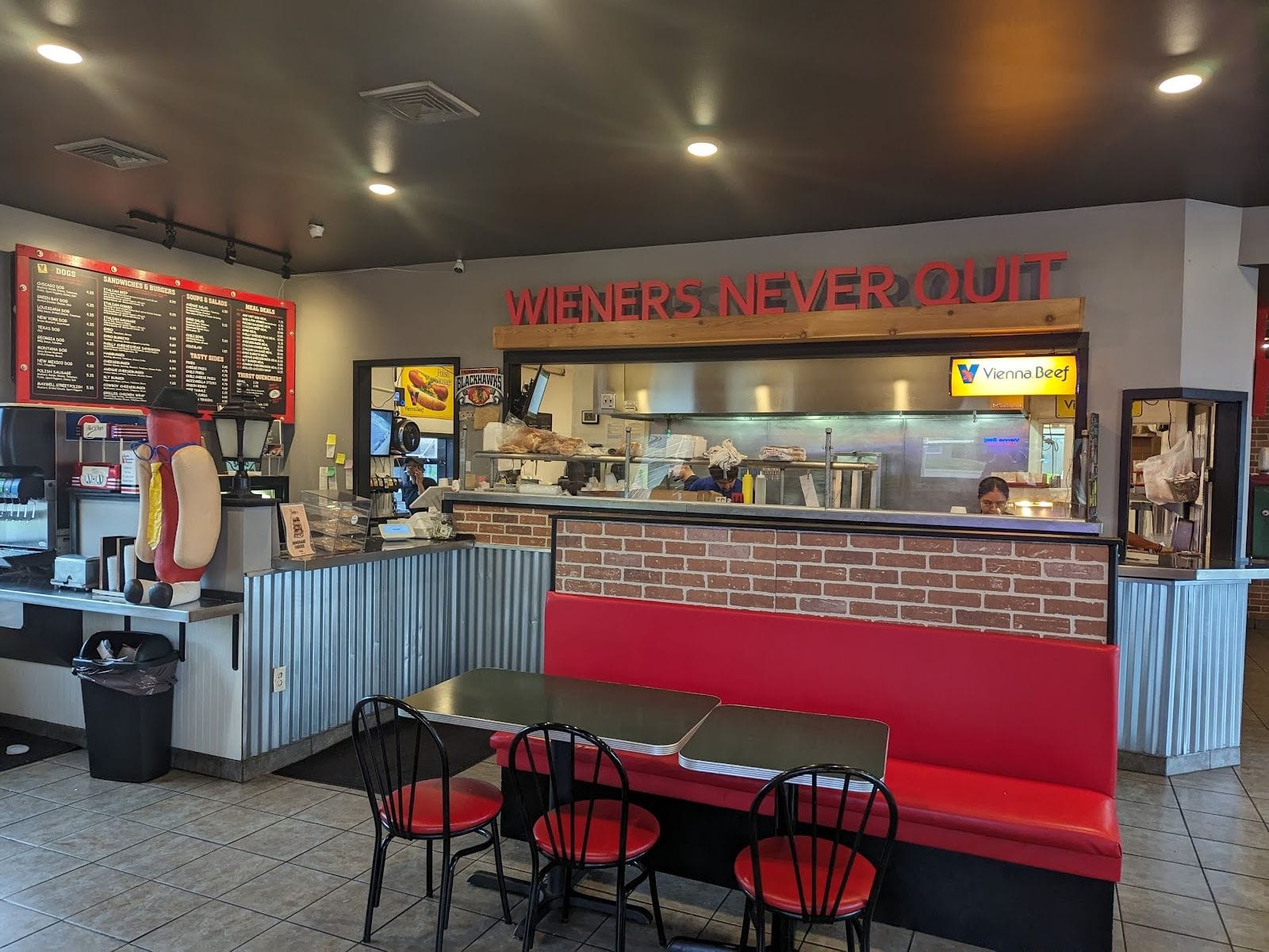 Interior of a hot dog restaurant with red seating, menu boards, and a hot dog statue by the counter.