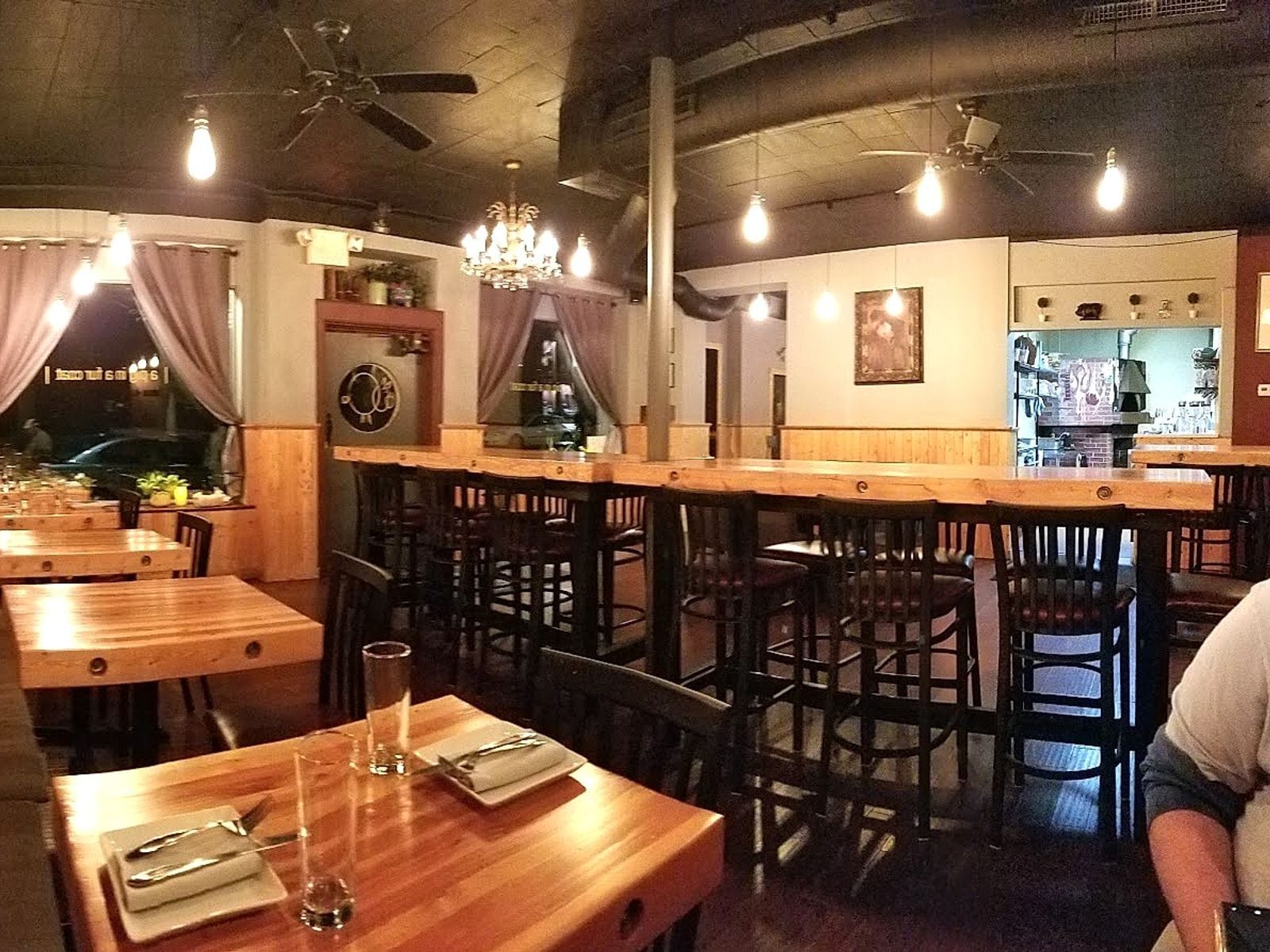 Warmly lit restaurant interior with wooden tables, high communal seating, and soft draped curtains.