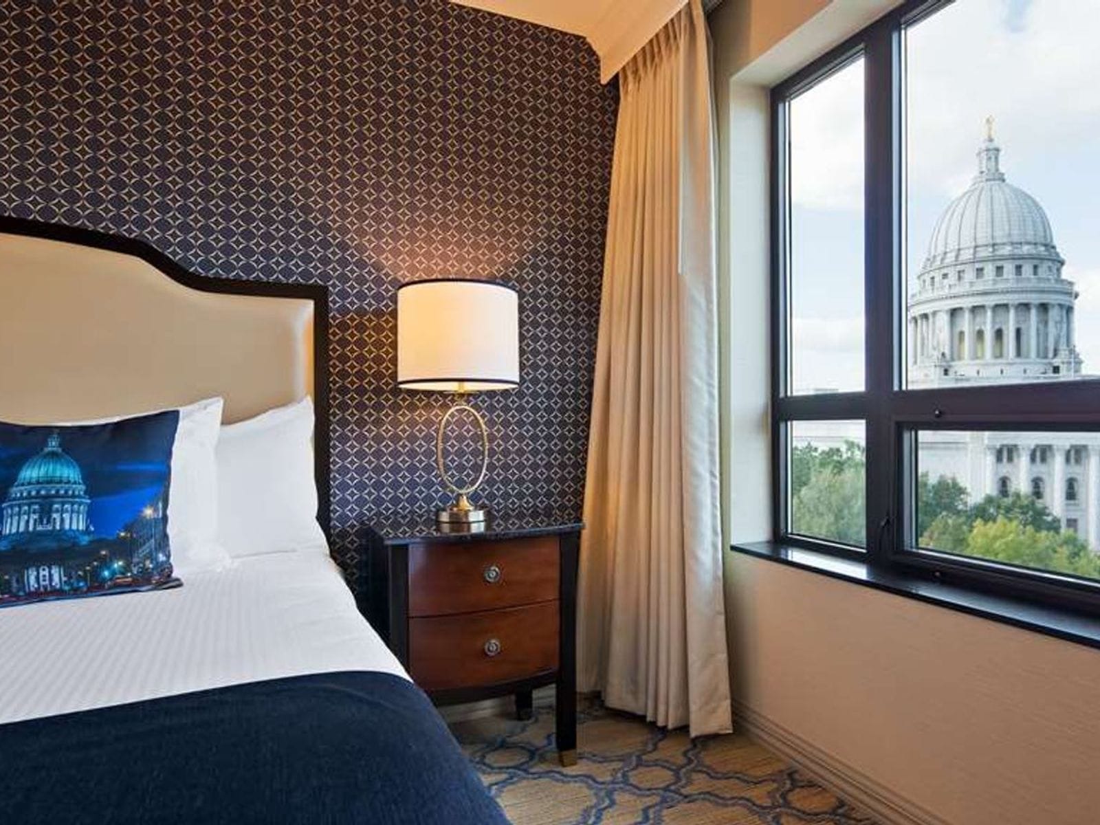 Modern hotel room with patterned wall, plush bed, lamp, and large window overlooking a domed capitol building.