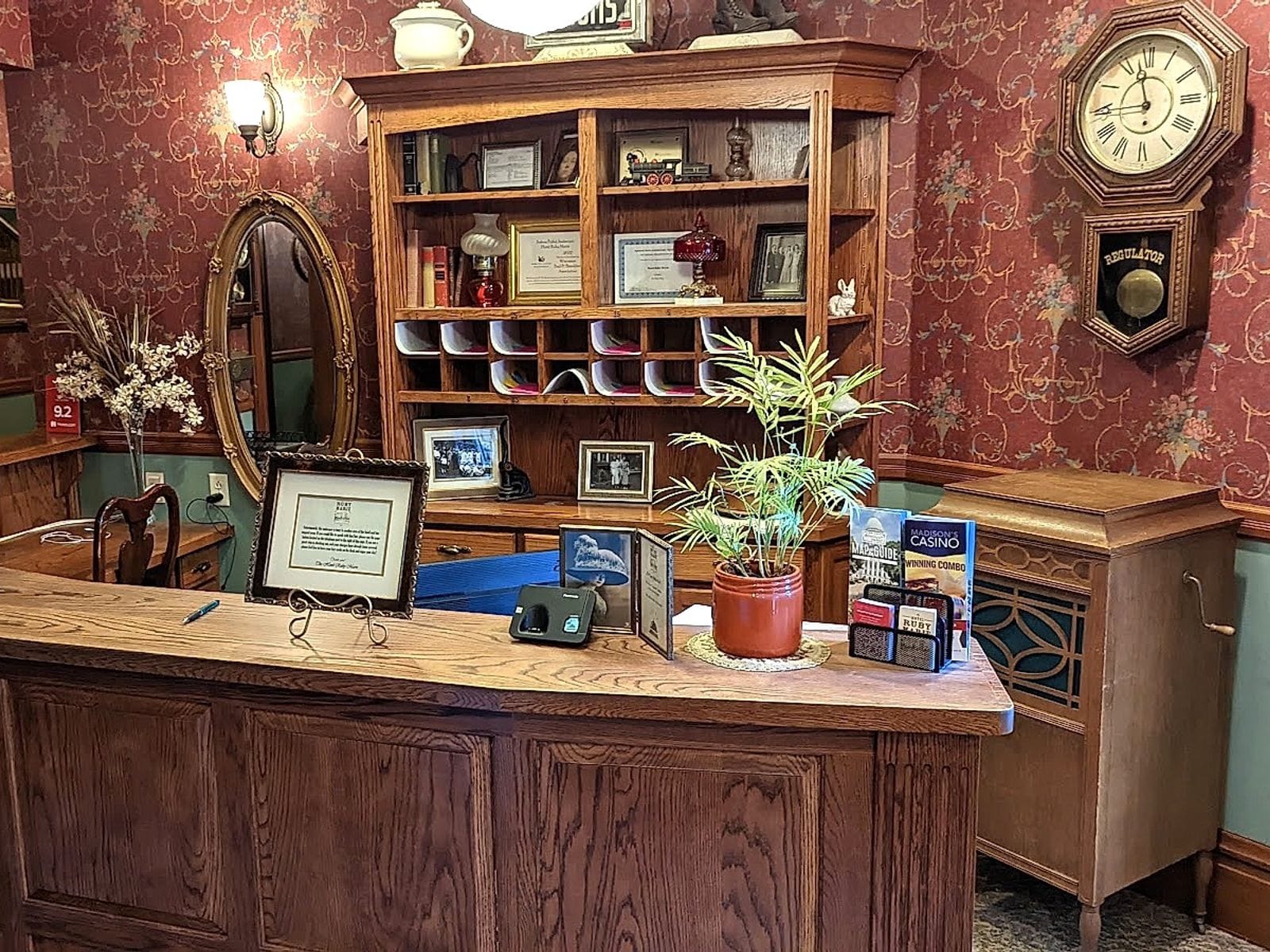 Ornate hotel reception desk with wood finishes, potted plant, brochures, vintage mirror, and wall clock.