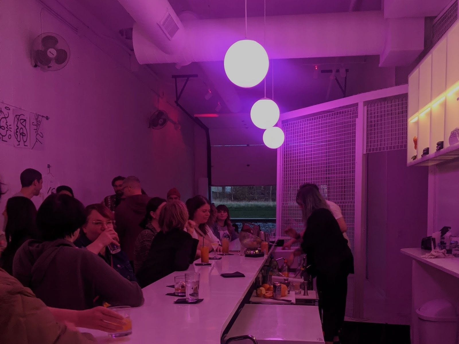 People sit at a long bar under glowing purple lights, with a bartender preparing drinks in a modern, cozy space.