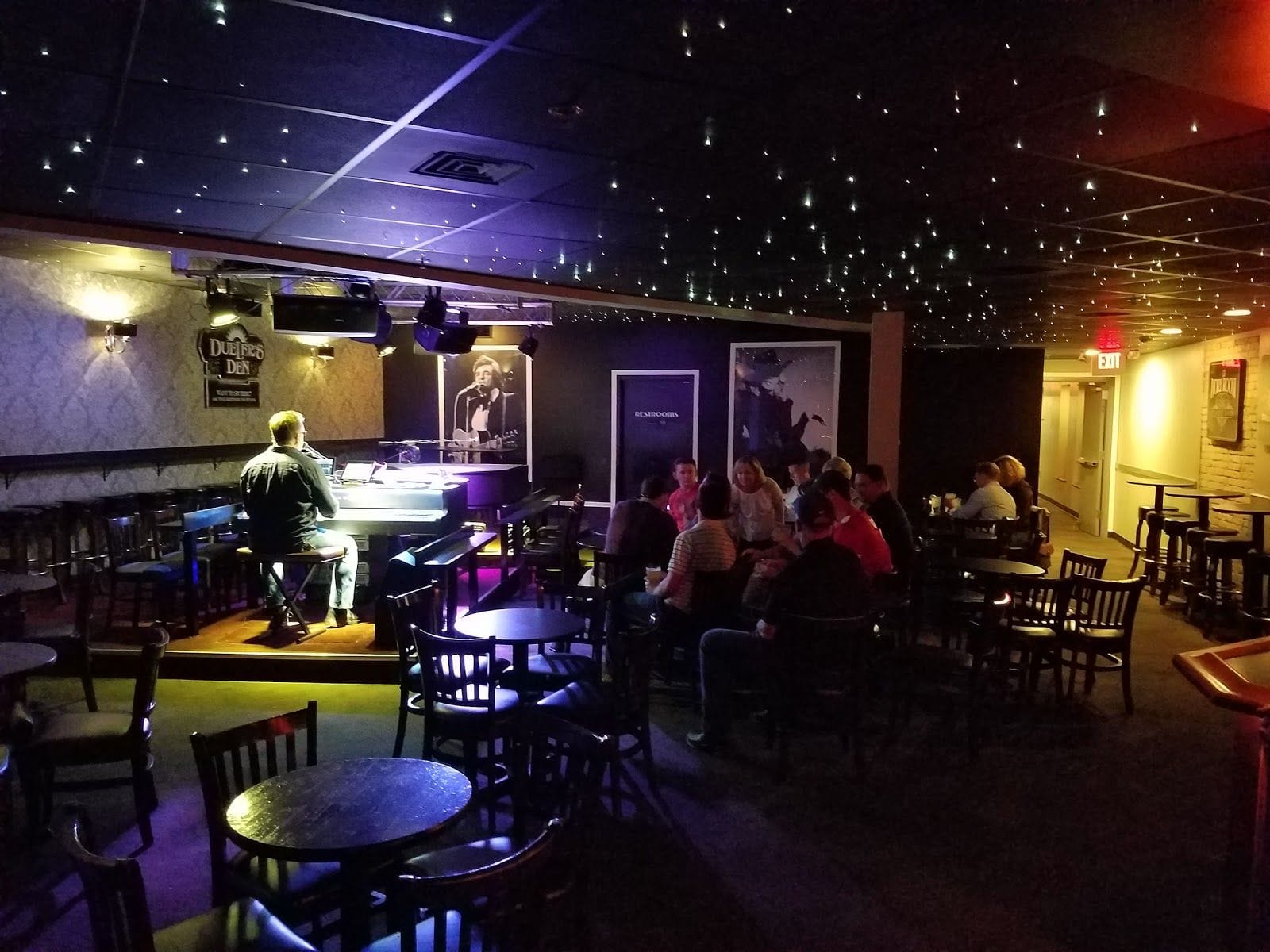 Man playing piano on a small stage in a dimly lit bar with people seated at tables under star-like ceiling lights.