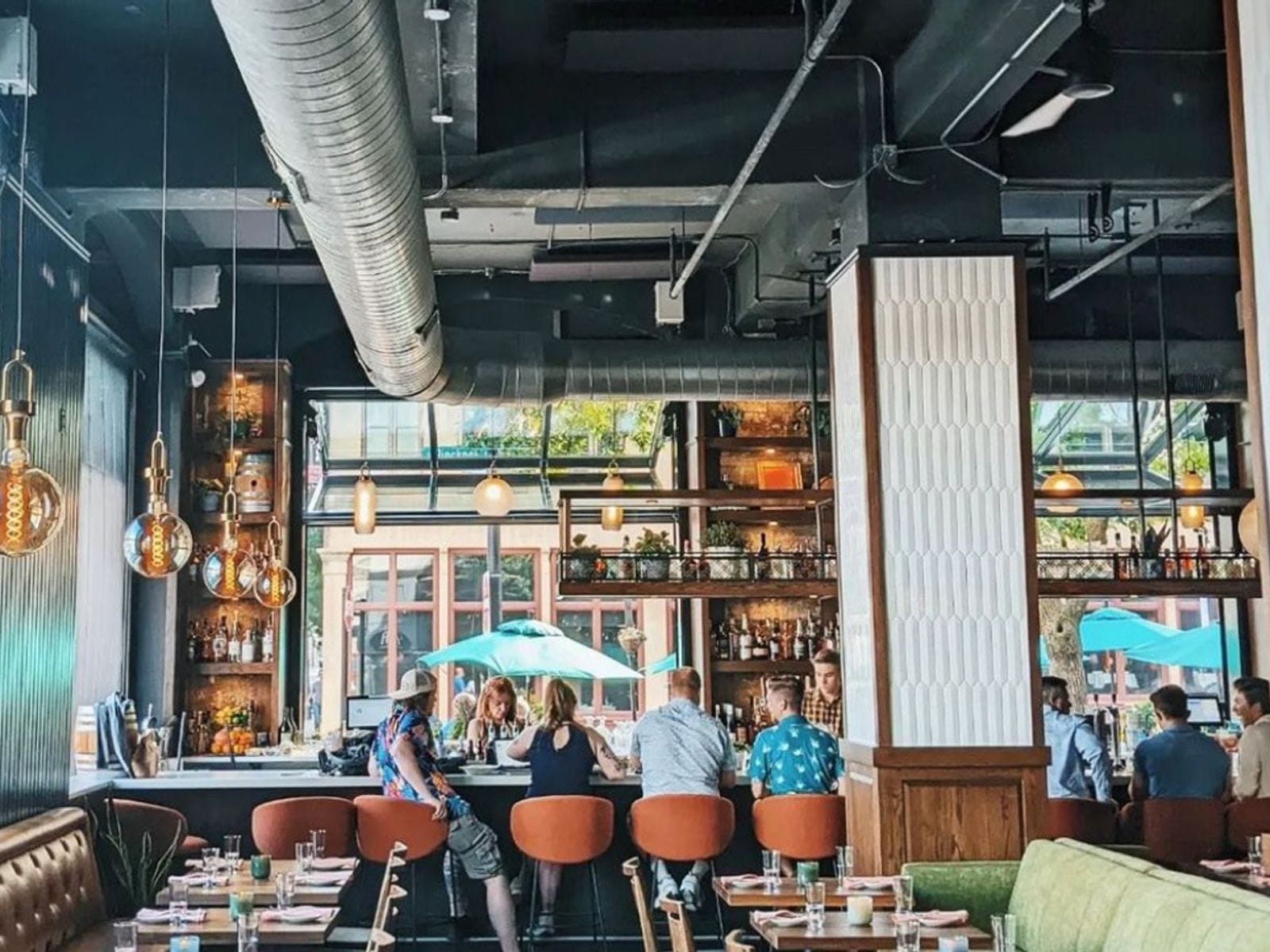 People sit at an industrial-style bar with orange stools, hanging lights, and large windows at Merchant Madison.