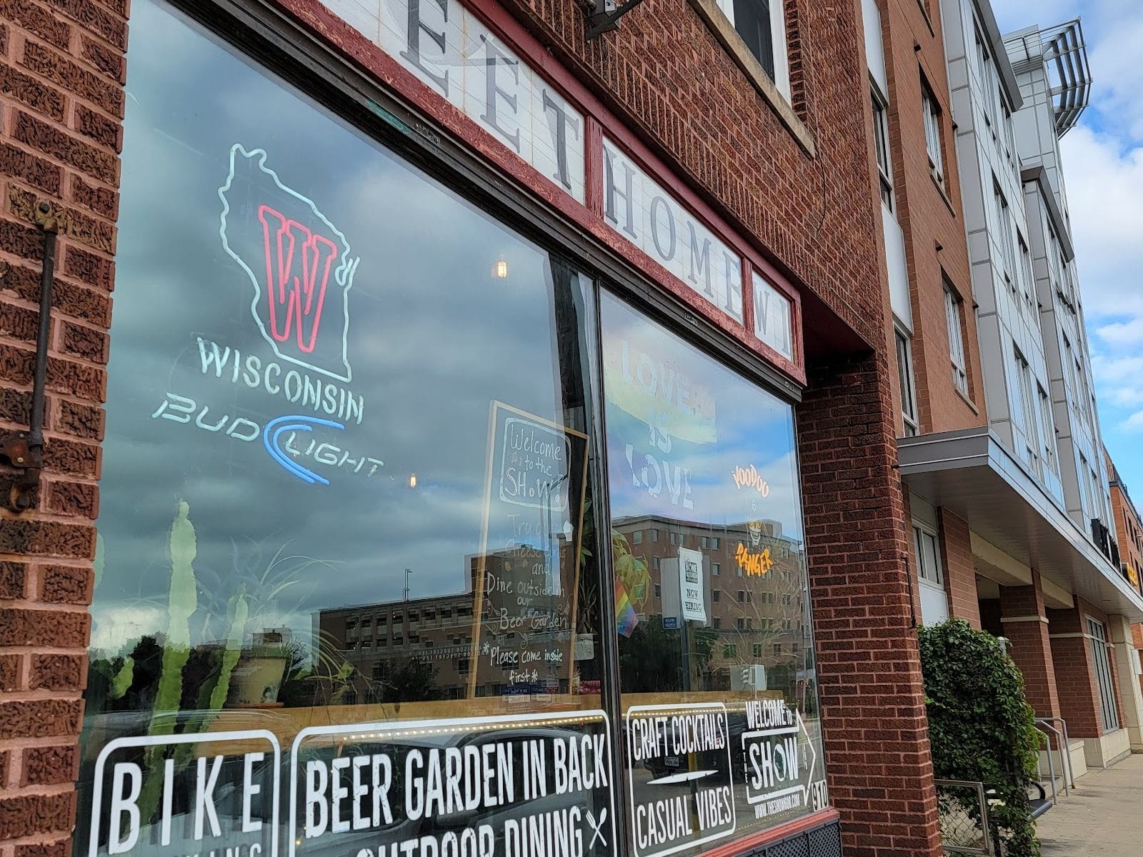 Brick building with large windows displaying neon signs, beer garden info, and reflections of the street.