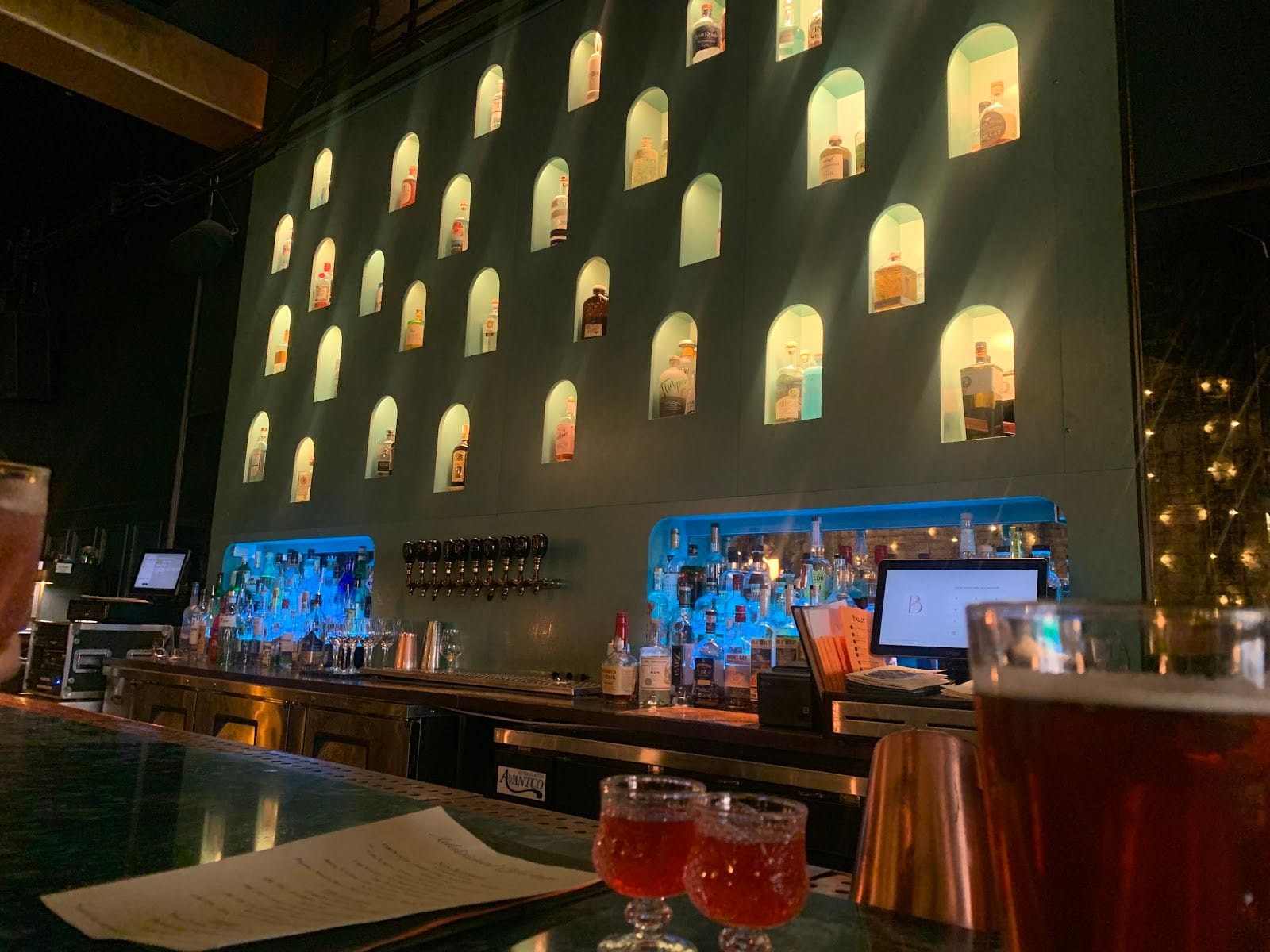 Backlit bar shelves with bottle alcoves, illuminated drinks, and a reflective countertop at The Botanist Social.
