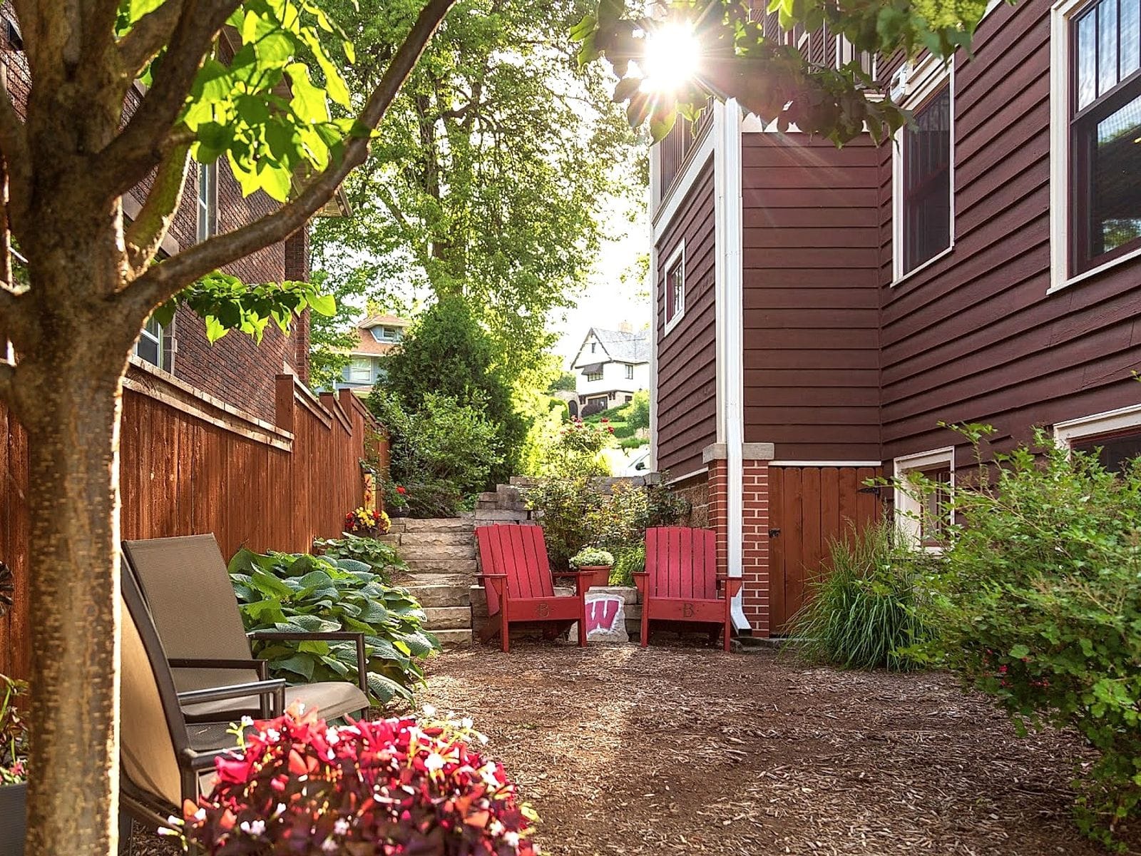 Sunlit garden patio with red Adirondack chairs, lush greenery, and brown siding of The Buckingham Inn.