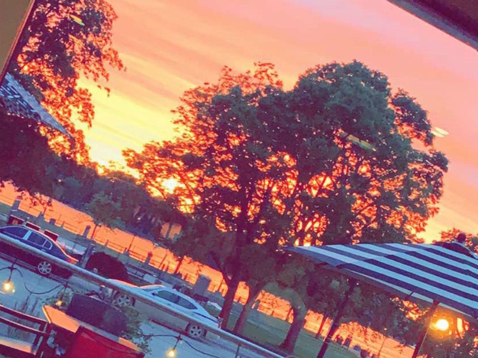 Orange-pink sunset over a river with silhouetted trees, striped patio umbrella, string lights and parked cars