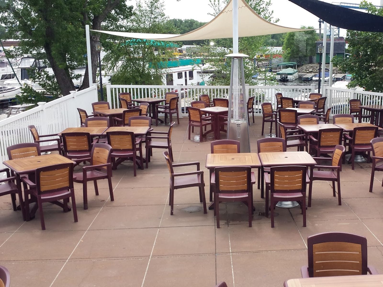 Riverside patio with wooden tables, maroon chairs, white railing and boats docked in background