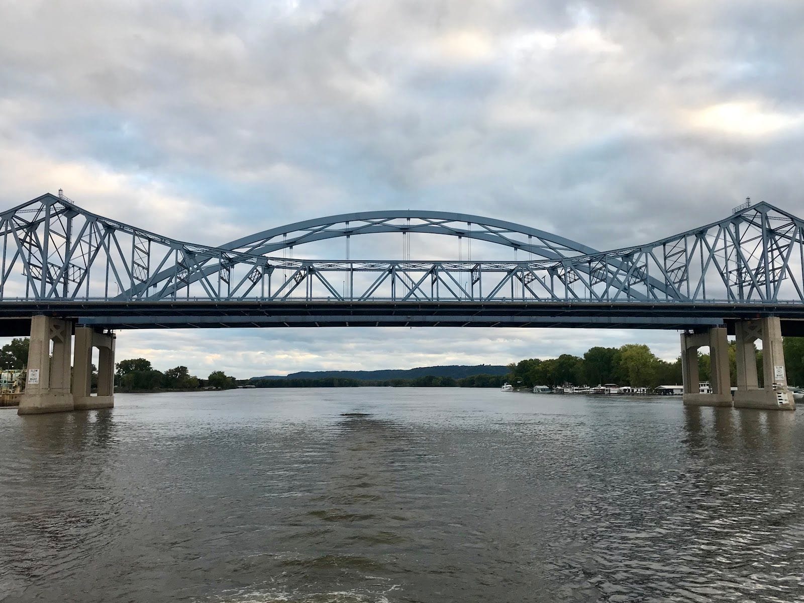 Steel truss arch bridge spanning a wide river under an overcast sky, concrete piers and tree-lined banks visible