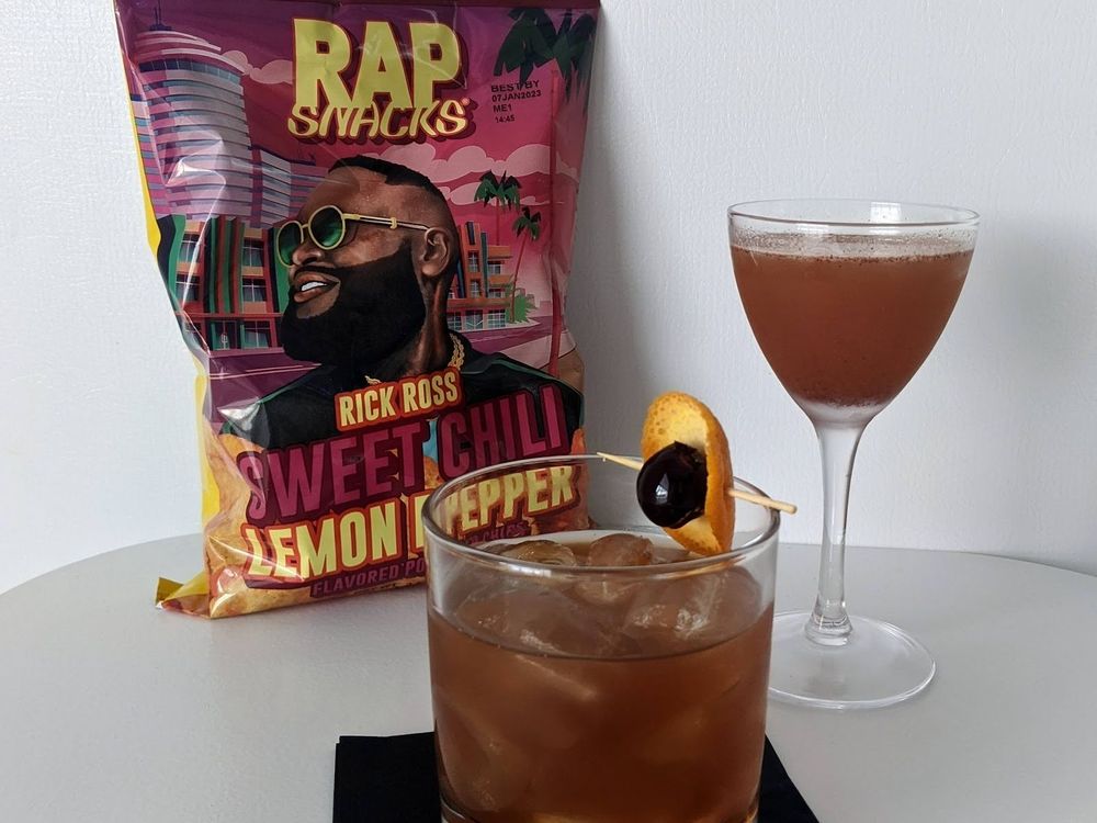 Two cocktails sit in front of a bag of Rap Snacks chips with a bold illustrated portrait on the packaging.
