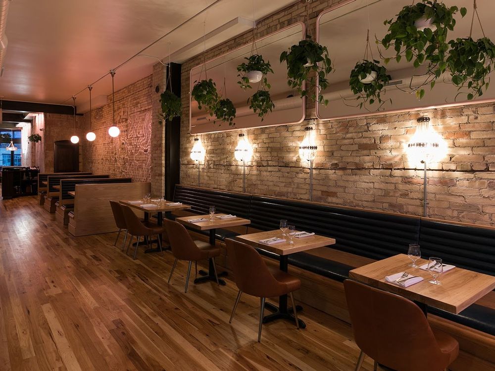 Cozy restaurant interior with exposed brick walls, wood tables, leather chairs, and hanging plants.