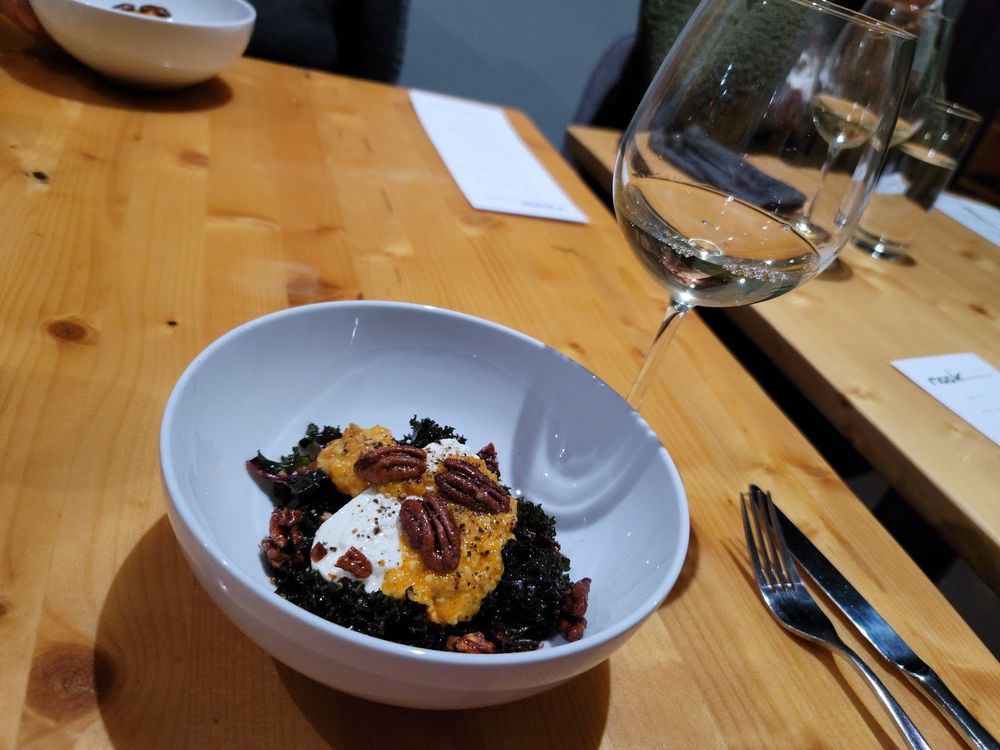 White bowl with salad, pecans, and cheese next to a glass of white wine on a wooden table.