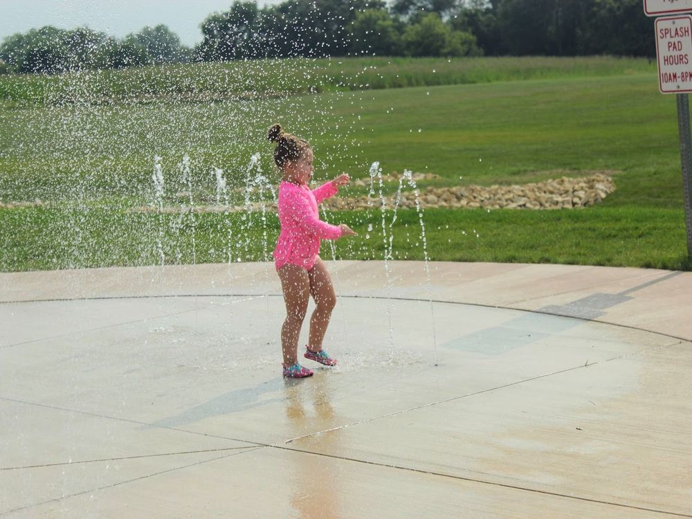 Young child in a bright pink swimsuit plays in arcing splash pad fountains on a sunny park day.