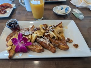 Golden pancakes topped with bananas, nuts, and a purple flower, served with syrup and orange juice.