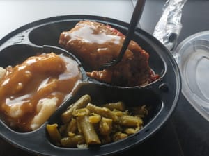 Black tray with meatloaf topped with gravy, mashed potatoes with brown gravy, and green beans.