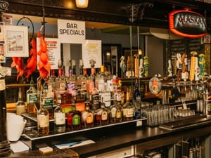 Assorted liquor bottles and beer taps line a mirrored bar counter with bar specials sign and glassware.