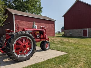 Vintage red Farmall tractor parked on concrete beside red barns and green grass at Schumacher Farm Park.