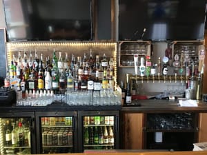 Bar counter with assorted liquor bottles, glassware, beer taps, and beverage coolers beneath two large TVs.