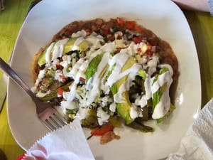 Mexican tostada topped with avocado slices, diced vegetables, and drizzled with white sauce on a white plate.